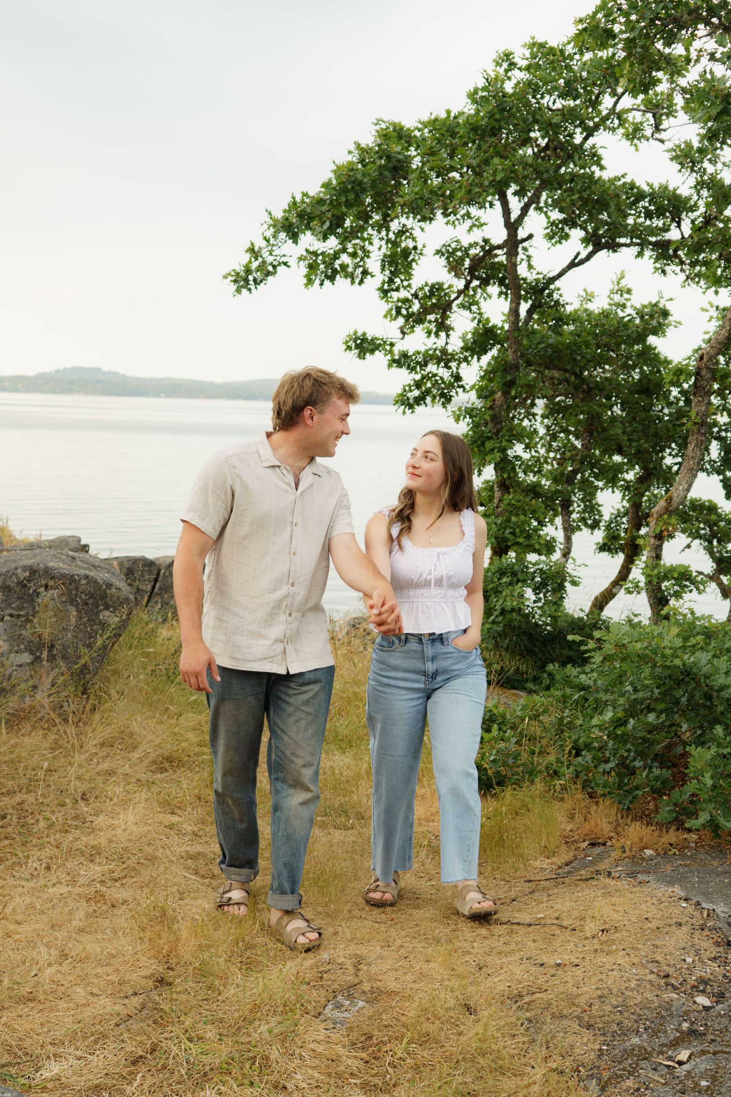 A young couple holds hands and smiles at each other while walking along a rocky shoreline with trees and water behind them.