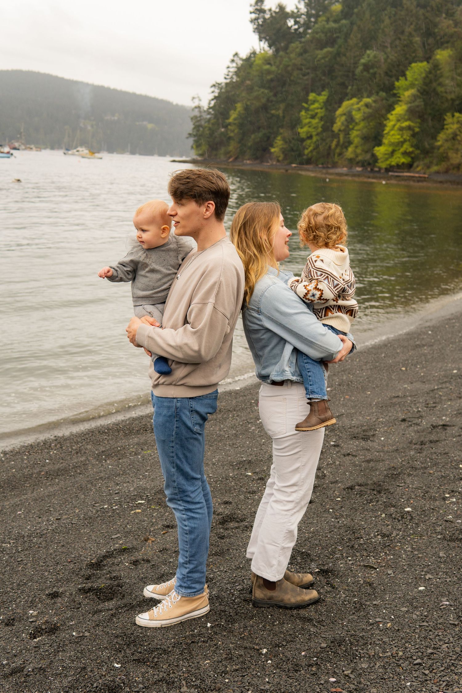 A young family stands back-to-back on a rocky beach, each parent holding a small child.