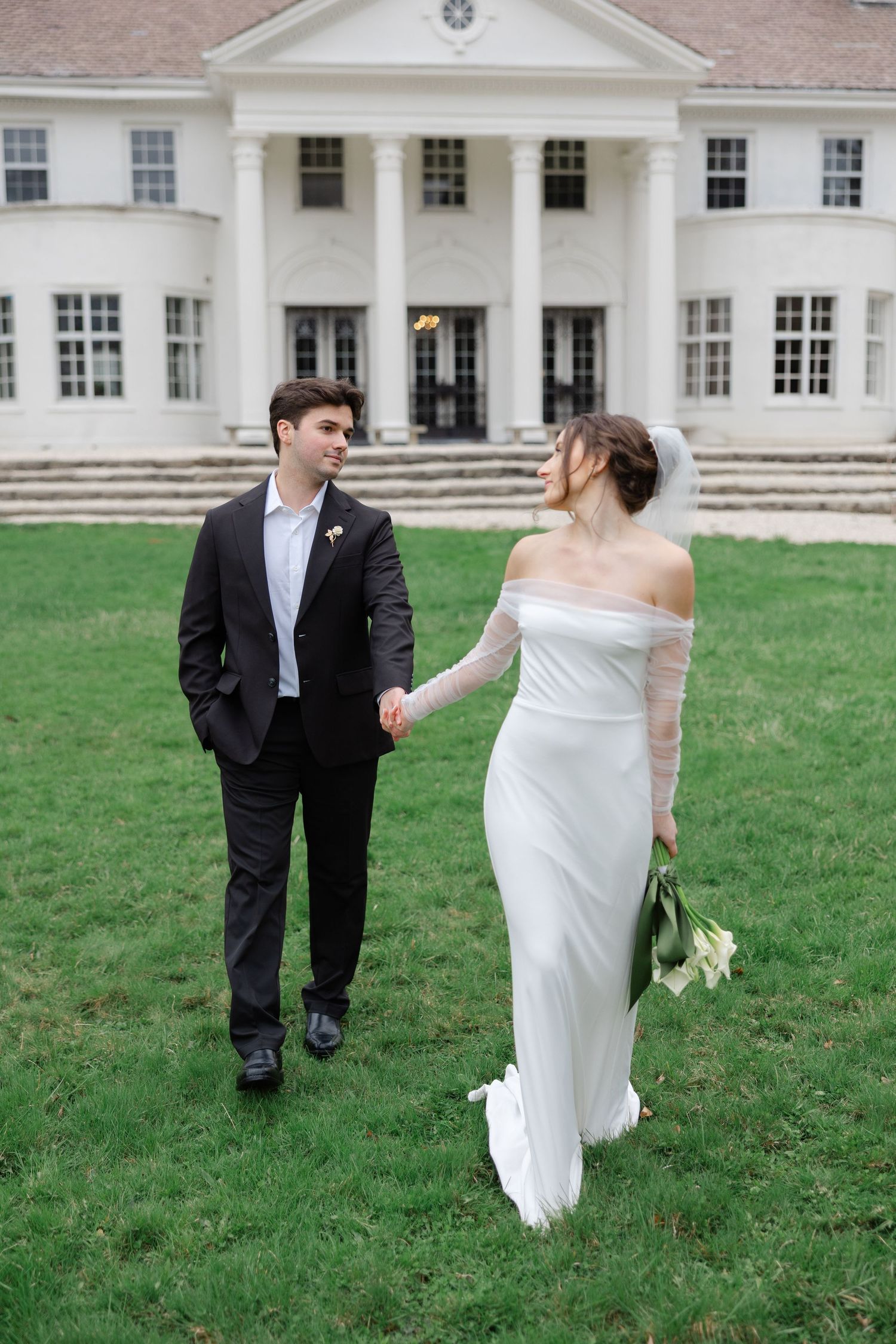 A bride and groom walk playfully across a green lawn in front of a grand white colonial-style mansion.