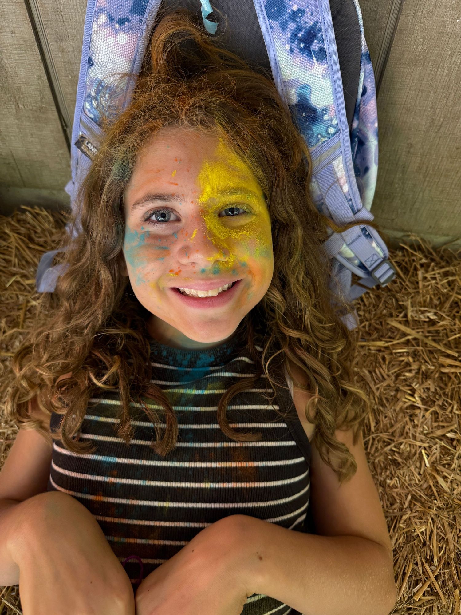 Smiling girl with colorful face paint lies back on hay in a cheerful outdoor setting.