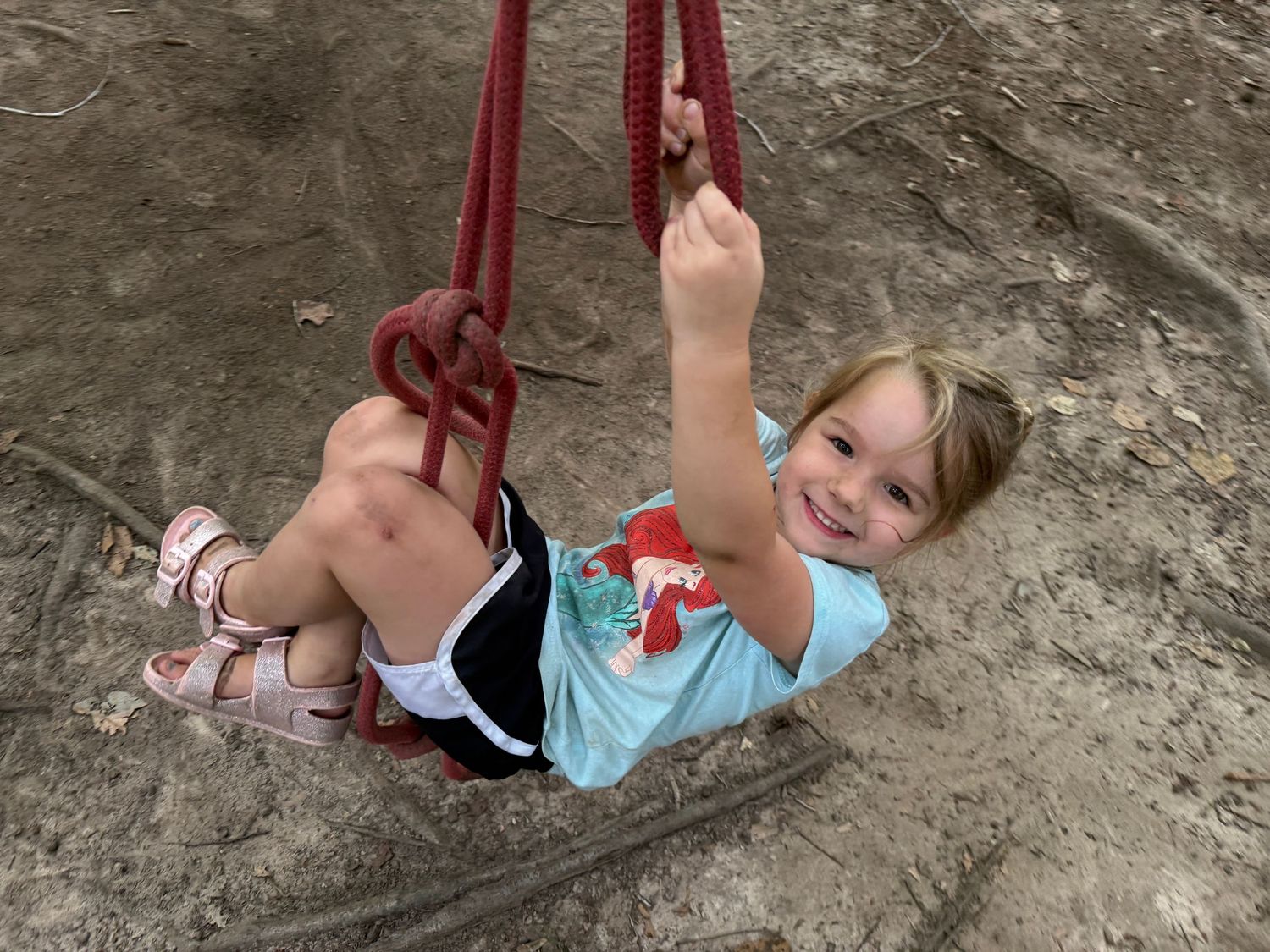 Smiling young girl swinging on a red rope swing at an outdoor playground.