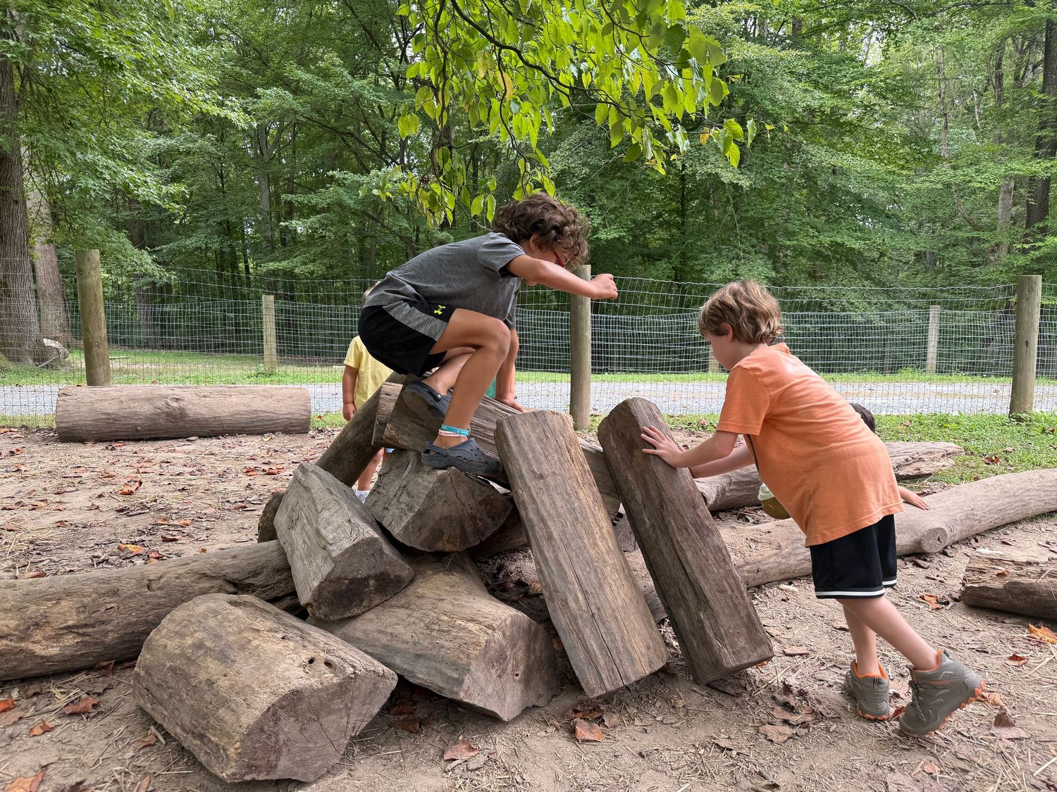 Two boys work together to climb and stack large wooden logs in an outdoor play area.
