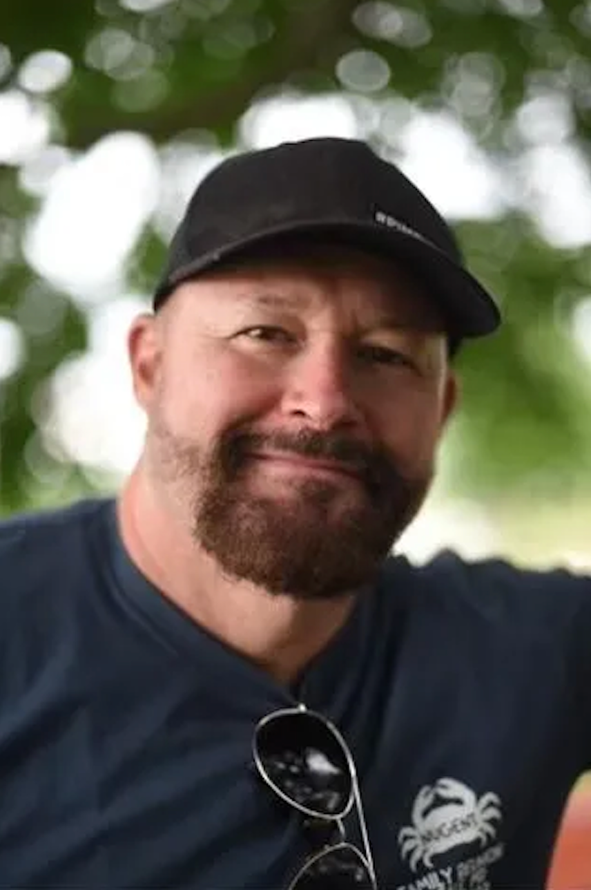 Smiling bearded man wearing a black cap and navy blue t-shirt outdoors with green trees in background.