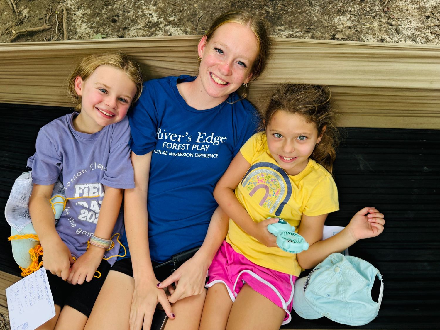Three smiling girls pose together wearing River's Edge Forest Play camp shirts outdoors.