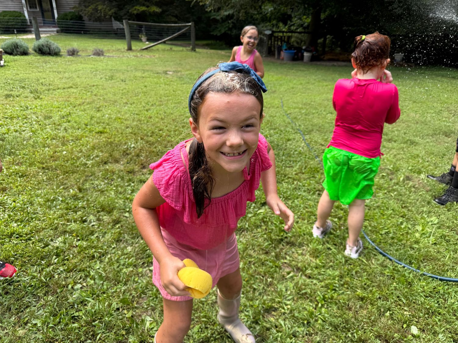 Smiling girl in pink holds a yellow water balloon during an outdoor water balloon fight.