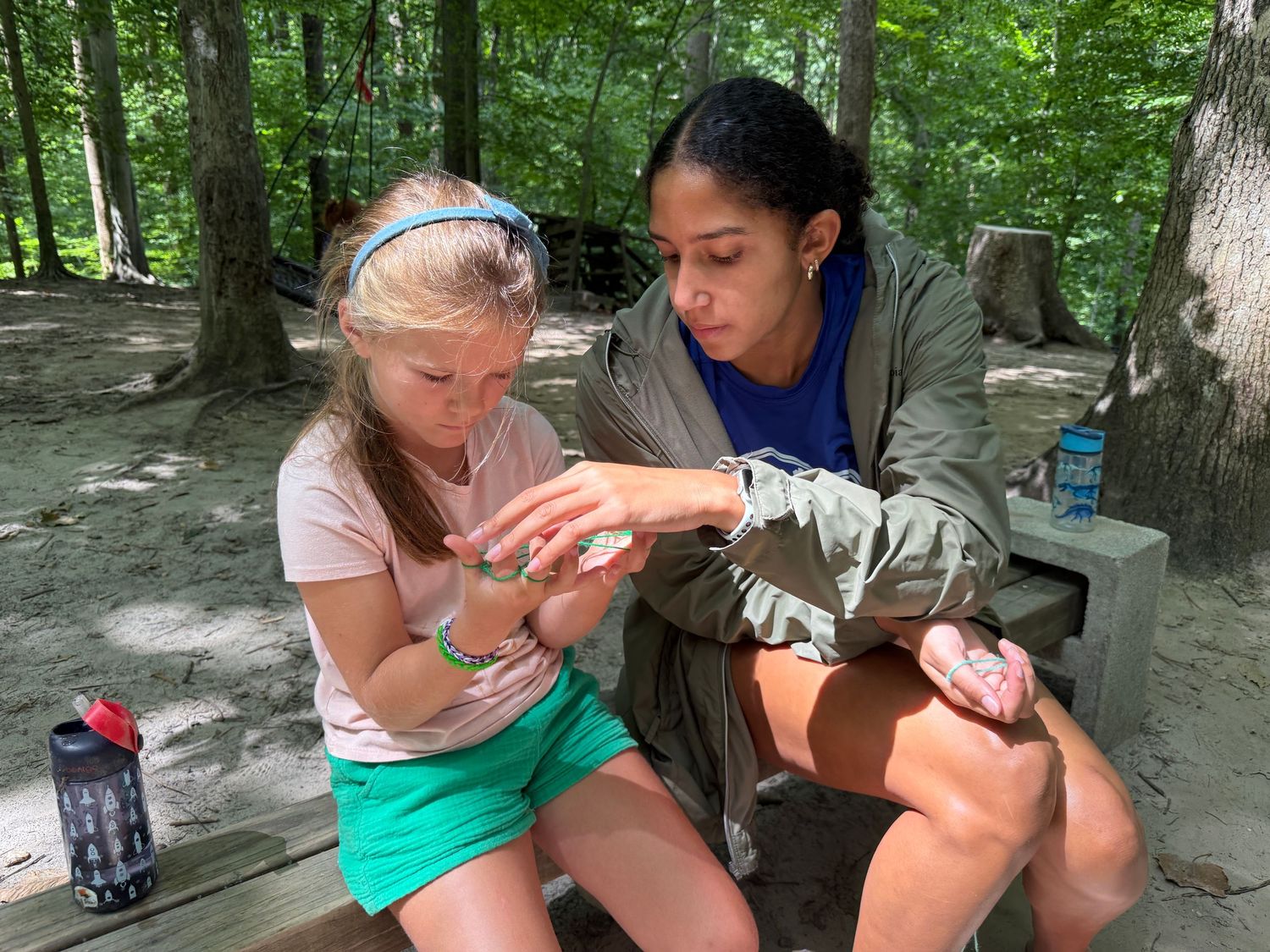 Adult woman helps a young girl examine something small in her hands at an outdoor bench.