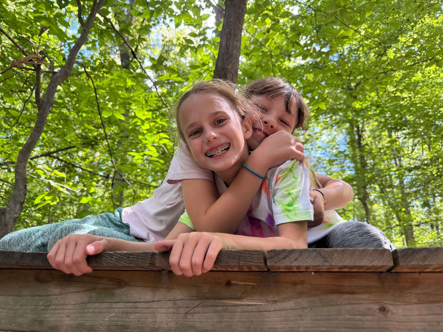 Two smiling girls lean over a wooden fence together in a lush green forest.