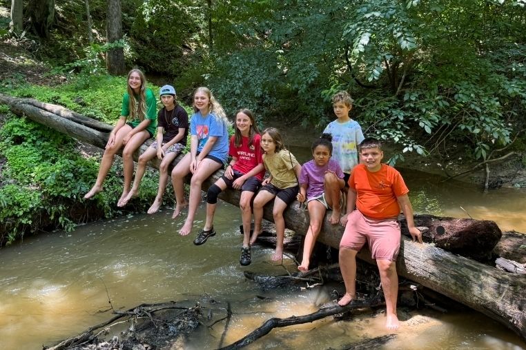 Seven children sit barefoot on a fallen log over a shallow creek surrounded by lush green trees.