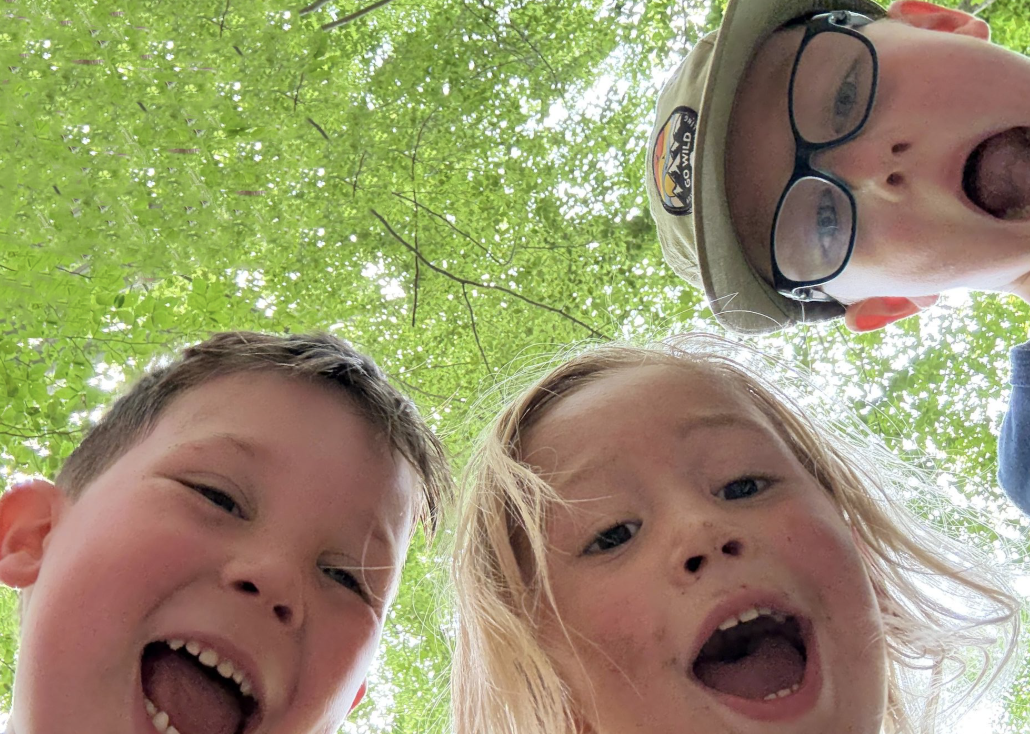 Three happy children laughing outdoors under lush green trees on a sunny day.