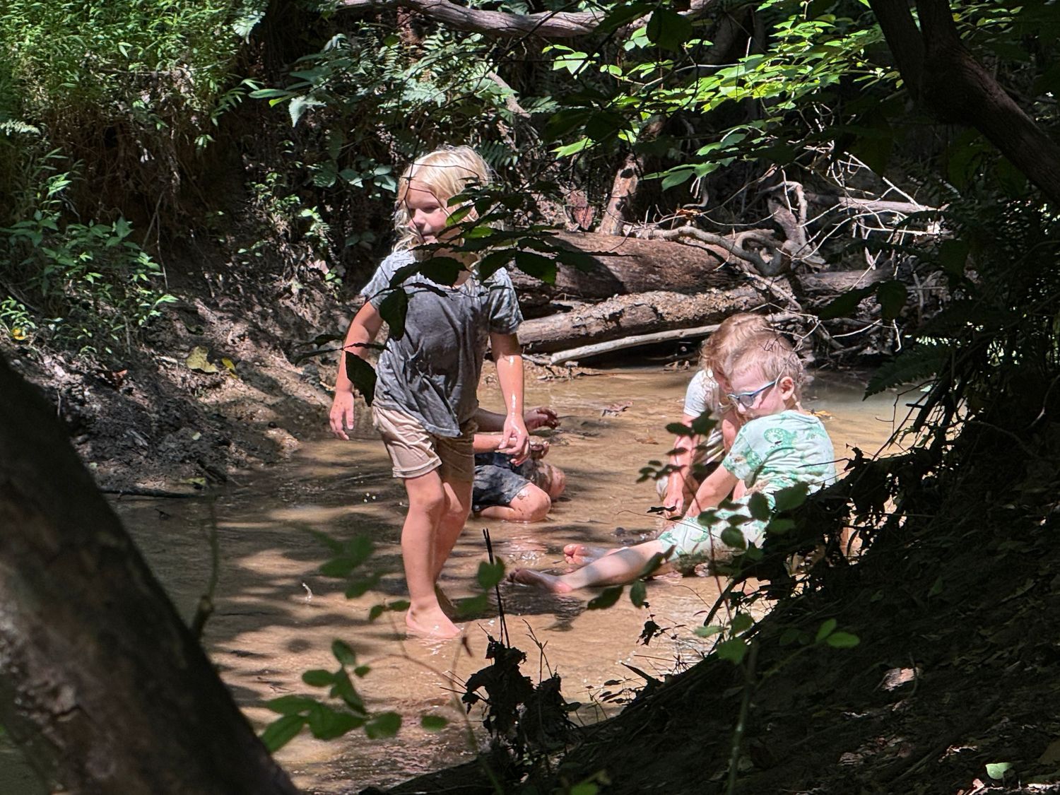 Two young children playing and splashing in a shallow muddy creek surrounded by nature.