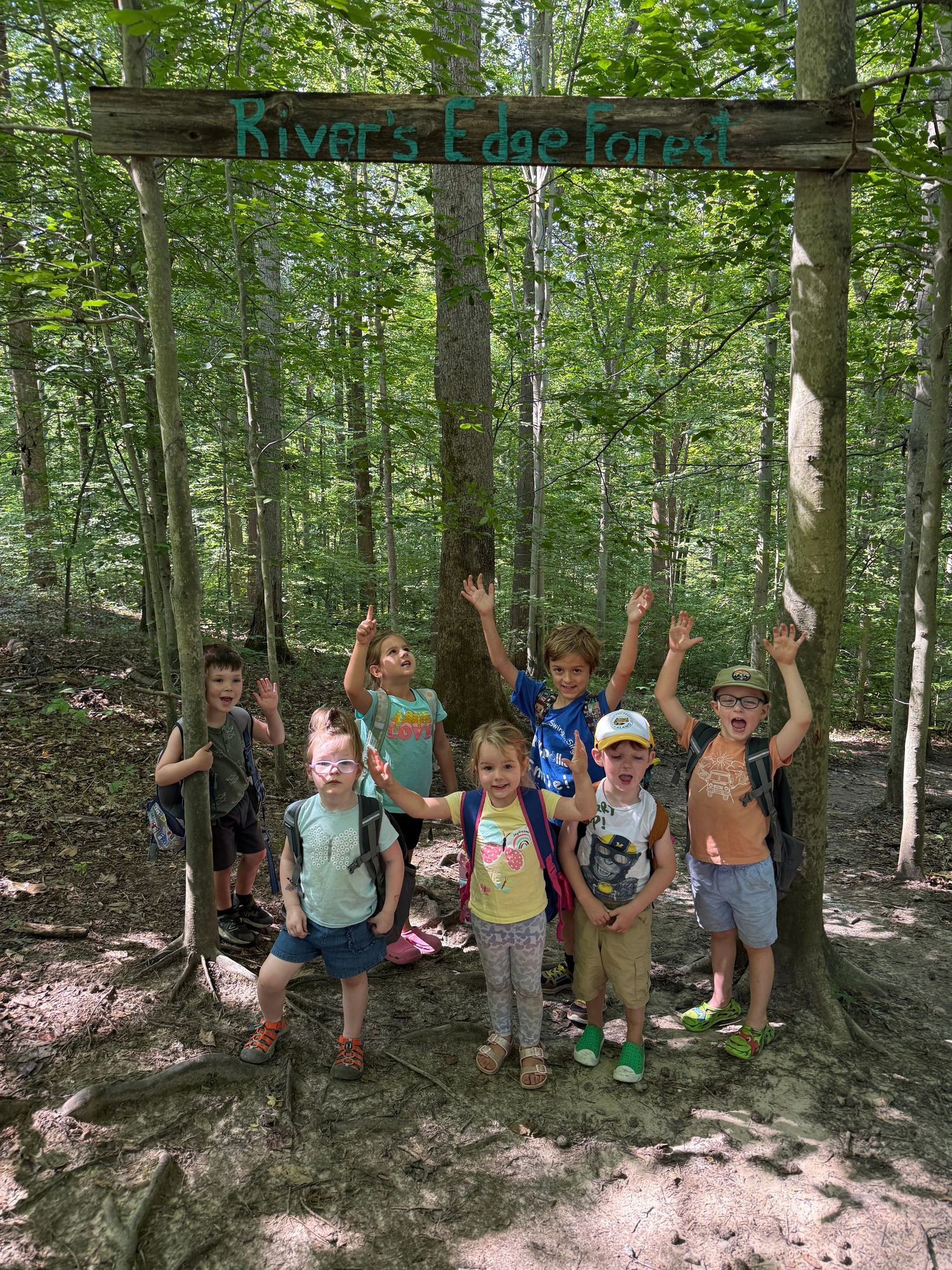 Children pose with raised hands under a wooden River's Edge Forest sign among lush green trees.