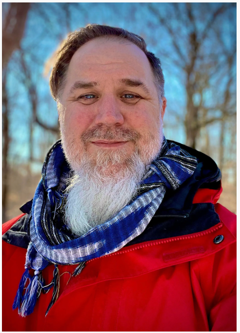 Middle-aged man with white beard wearing red jacket and blue scarf outdoors in winter.