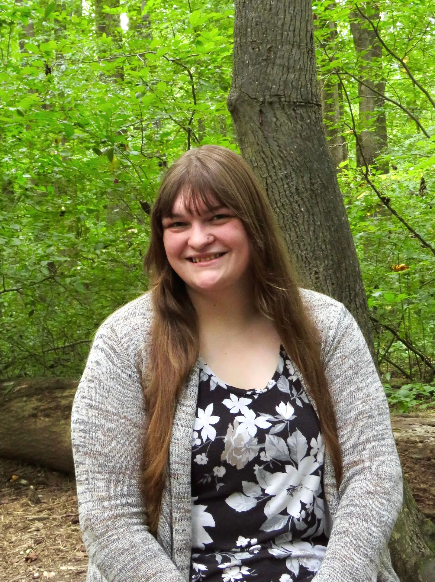 Young woman with long brown hair smiling in a forest wearing a gray cardigan and floral top.