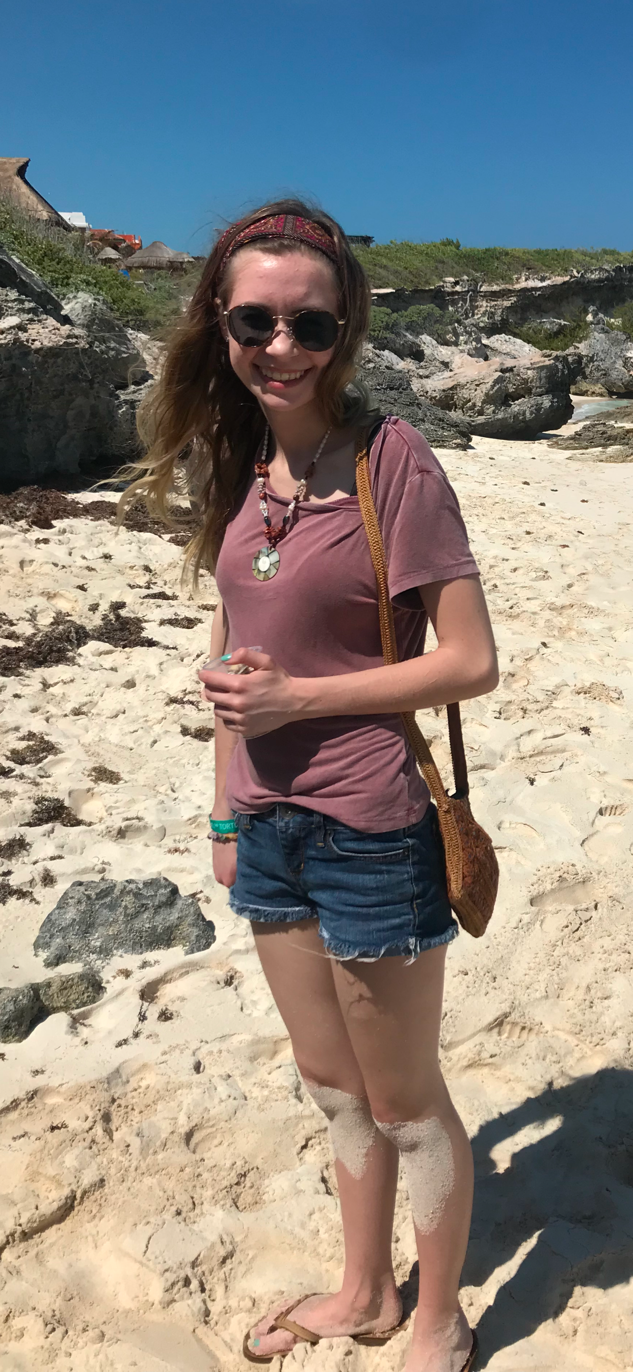Young woman smiling on sandy beach wearing denim shorts, pink tee, sunglasses, and crossbody bag on sunny day.