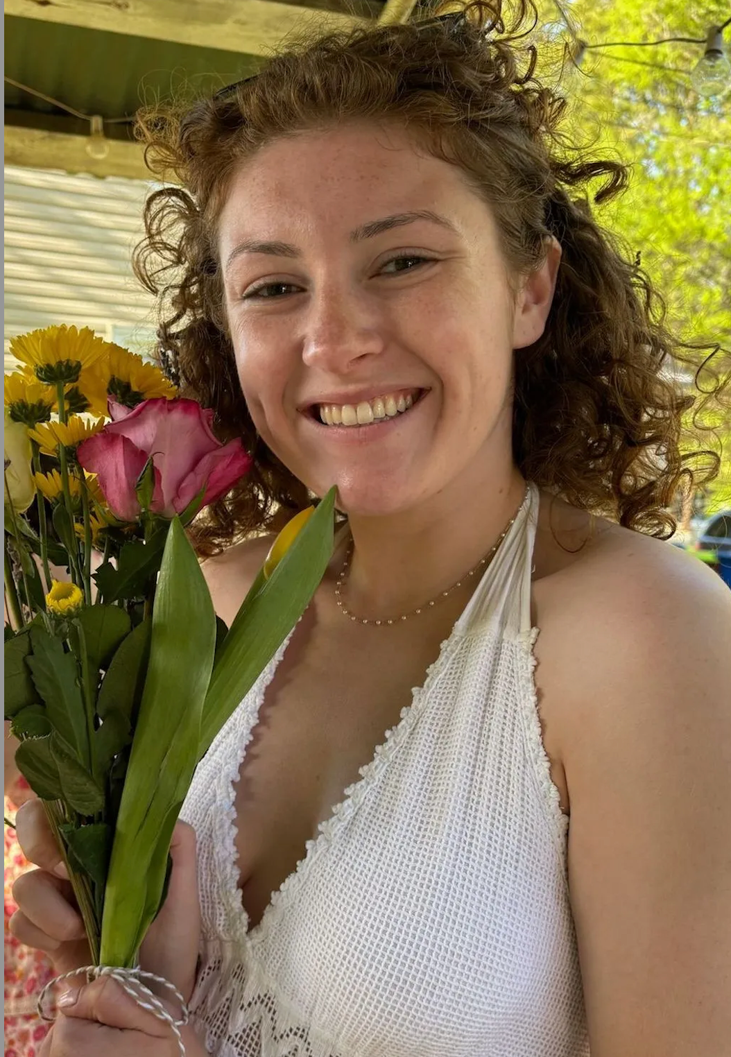 Young woman with curly red hair smiling while holding a colorful bouquet of flowers outdoors.