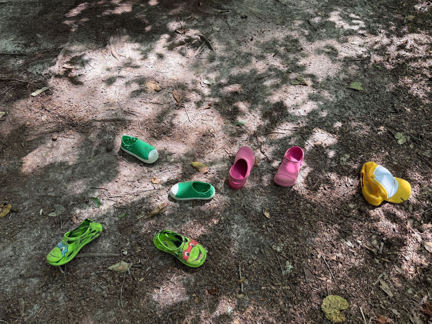 Colorful children's shoes left on the ground in dappled sunlight on a dirt path.