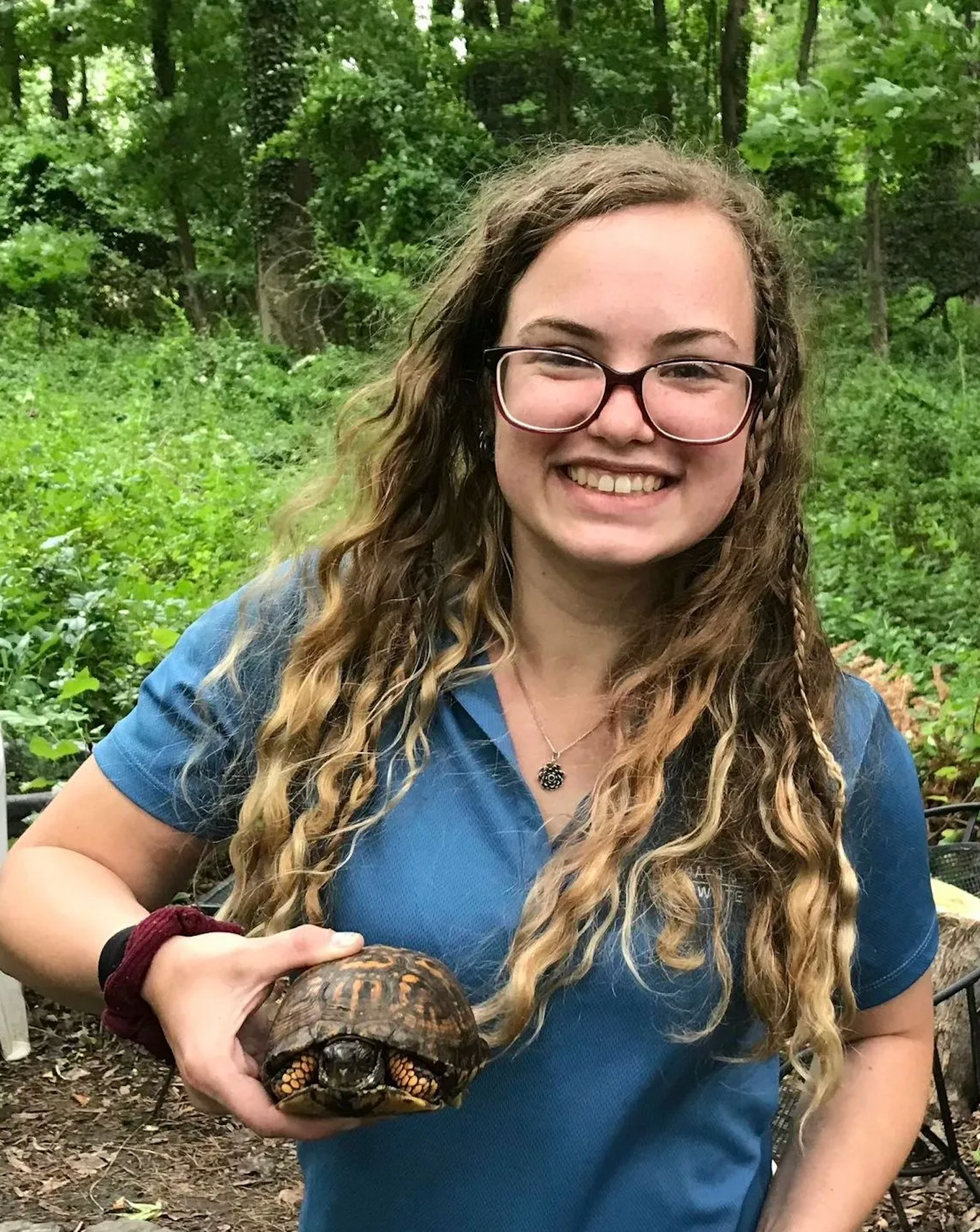 Smiling young woman with curly blonde hair holding a box turtle outdoors in a wooded area.
