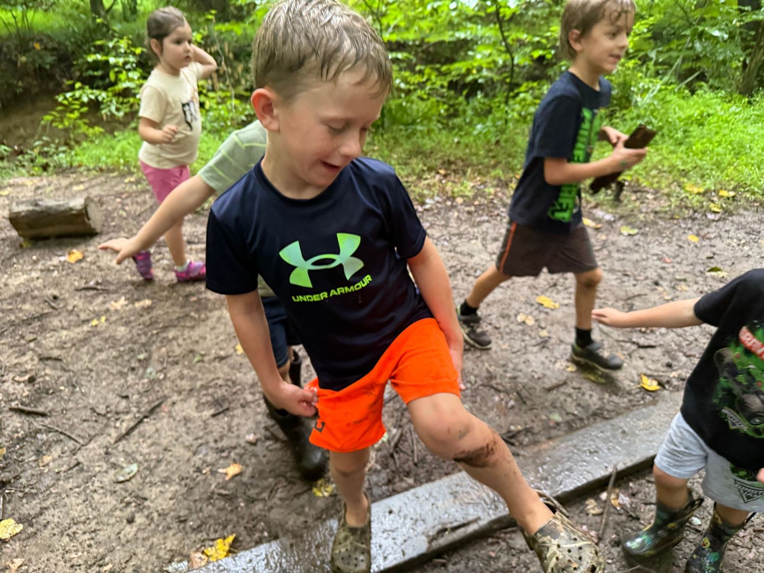 Young boy in Under Armour shirt runs through muddy terrain in a wooded outdoor area.