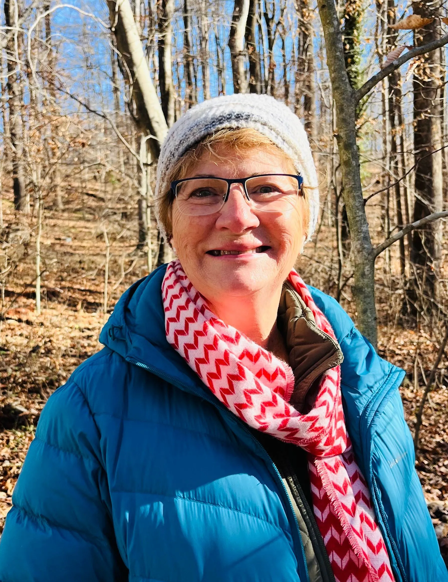 Older woman smiling outdoors in autumn woods wearing a teal jacket, red scarf, and gray beanie.
