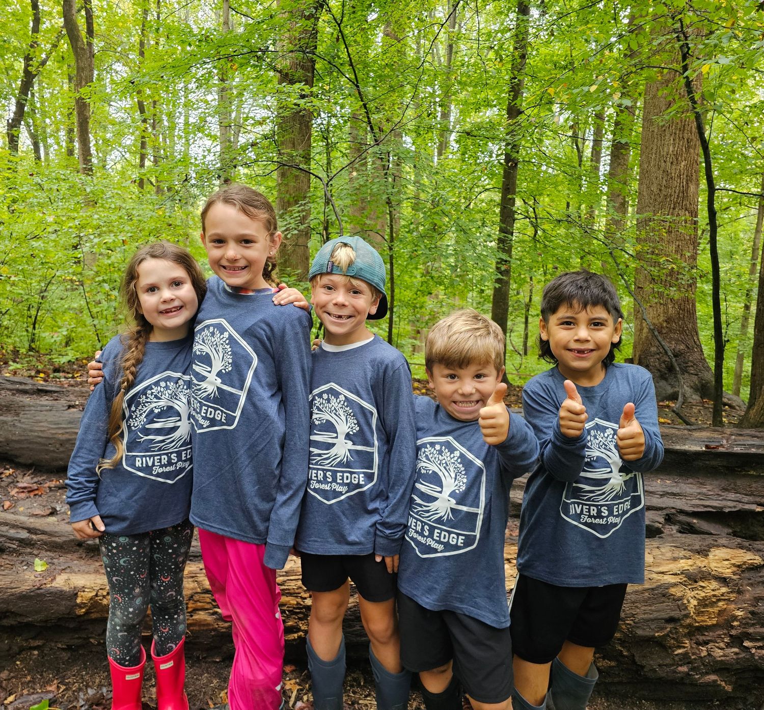 Five smiling children in matching River's Edge Forest Play shirts posing together in a green forest.