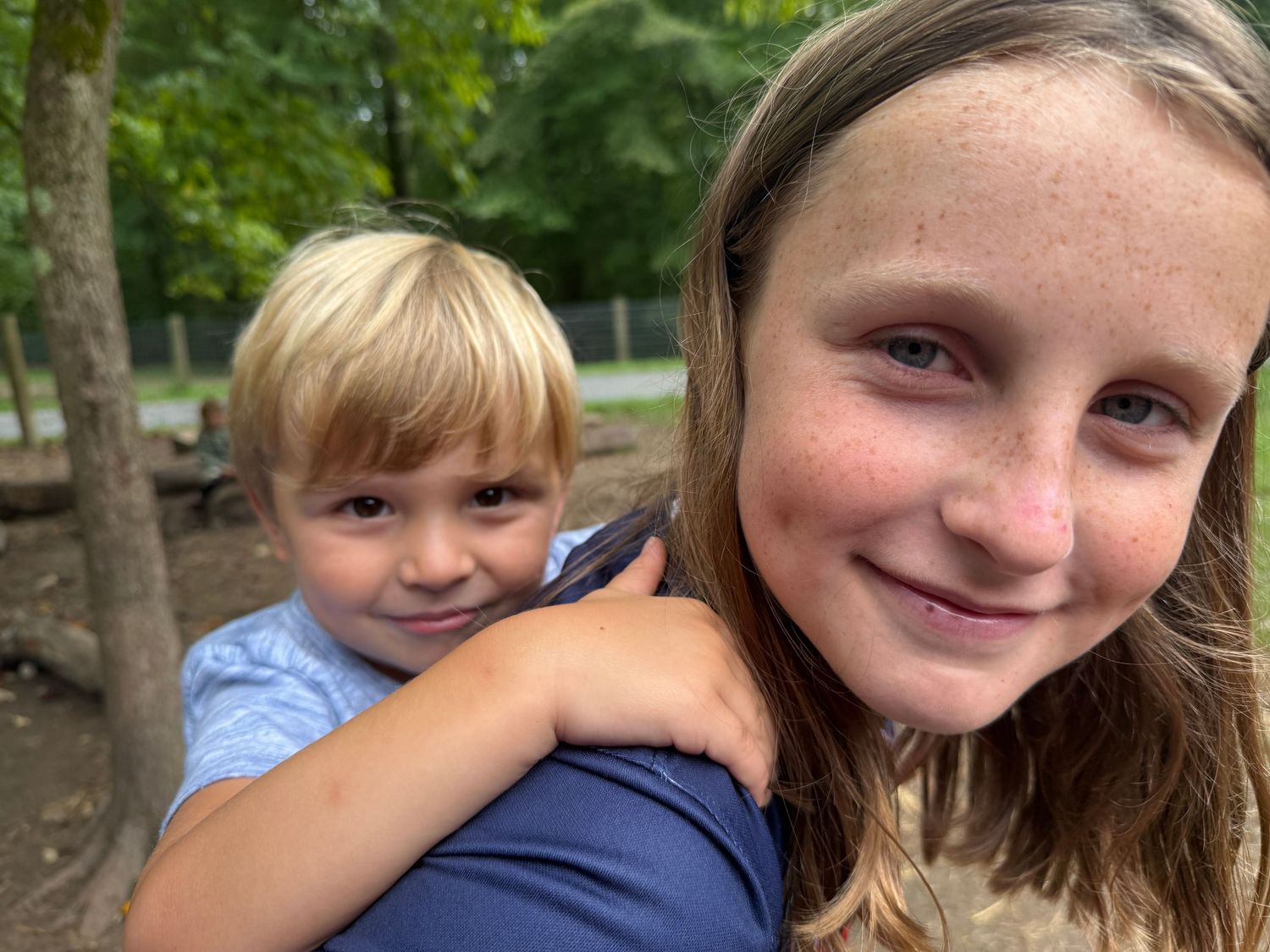Young girl smiles as a small blonde boy hugs her from behind outdoors near trees.