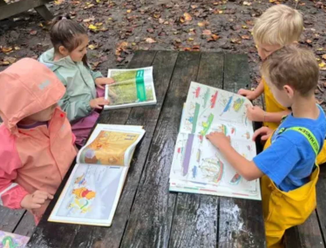 Children in rain gear studying nature books at a wet picnic table in the forest.