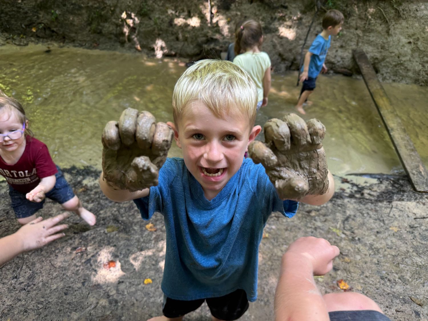Excited boy shows off muddy hands near a creek during outdoor nature exploration.
