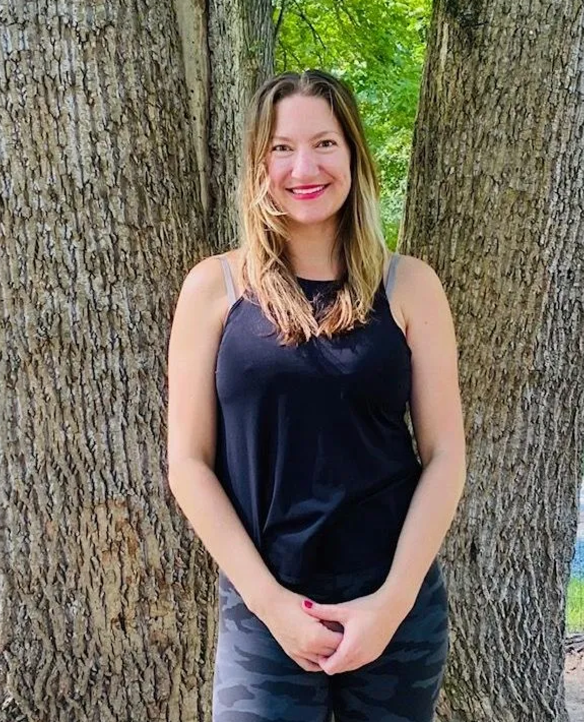 Woman smiling outdoors between two large trees, wearing a black tank top and patterned leggings.