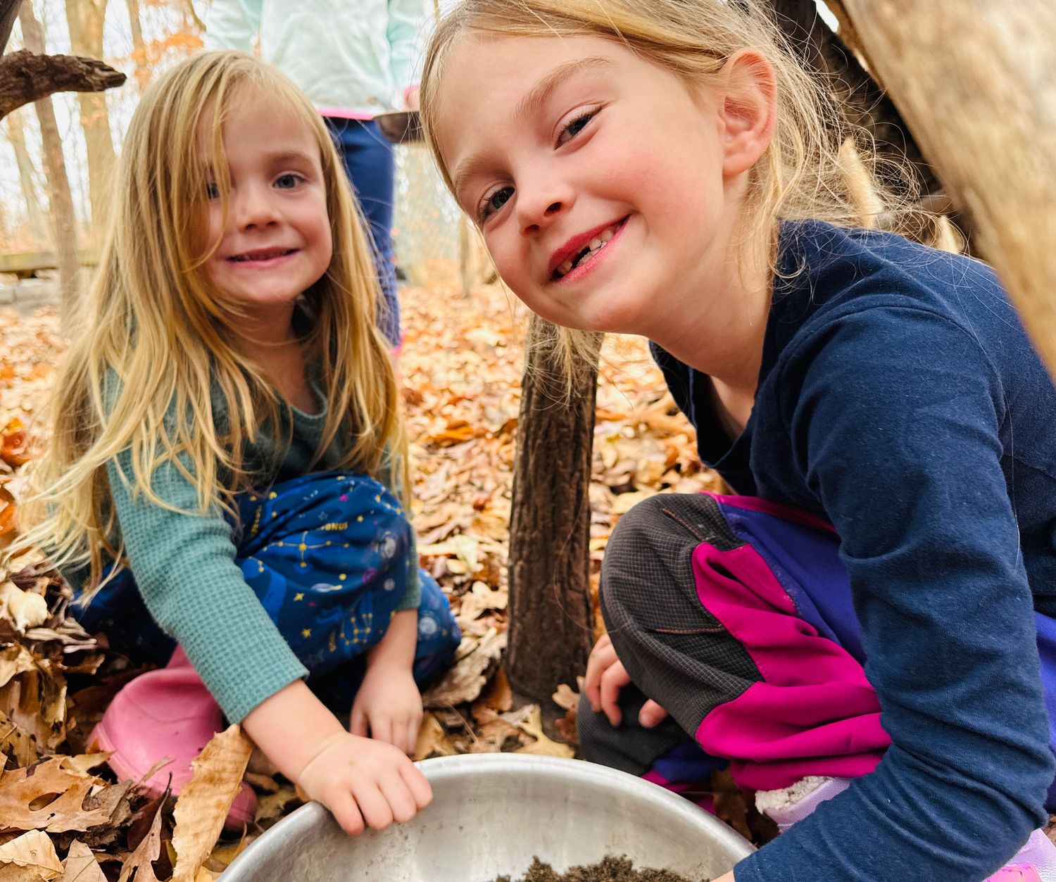 Two young girls playing outdoors in autumn leaves, exploring nature together with a metal bowl.