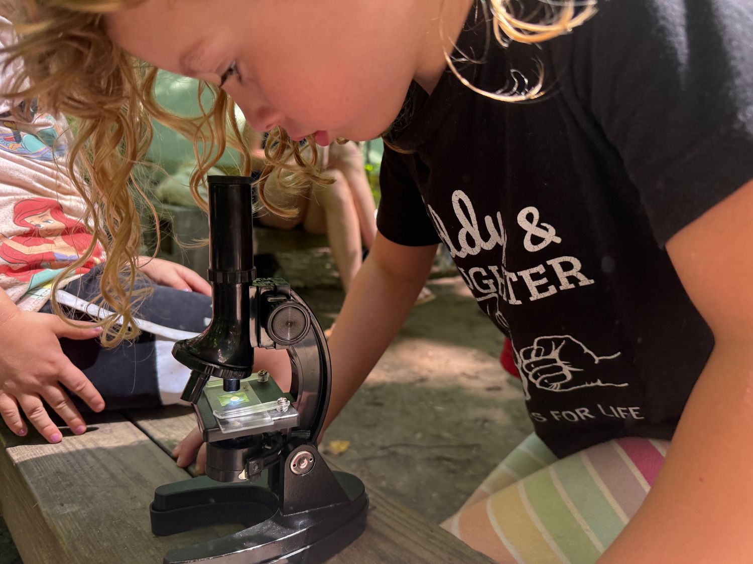 Young girl with curly hair looks through a microscope during outdoor science activity.