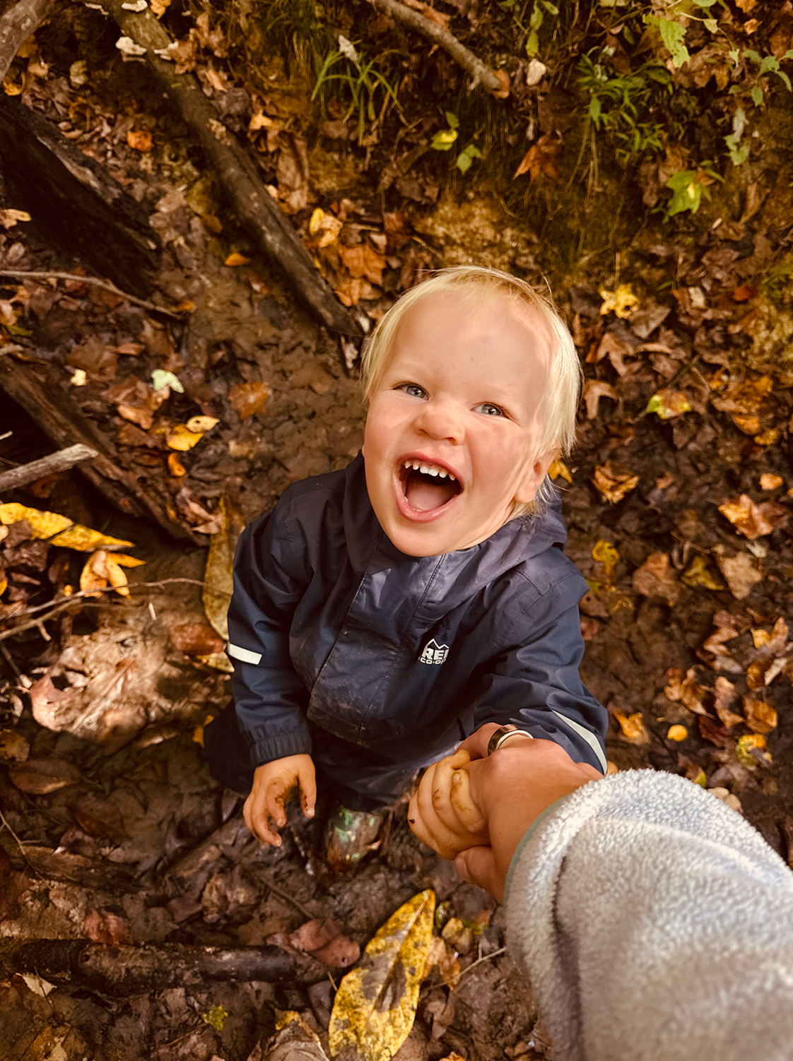 Joyful toddler with blonde hair laughing while playing in mud and autumn leaves outdoors.