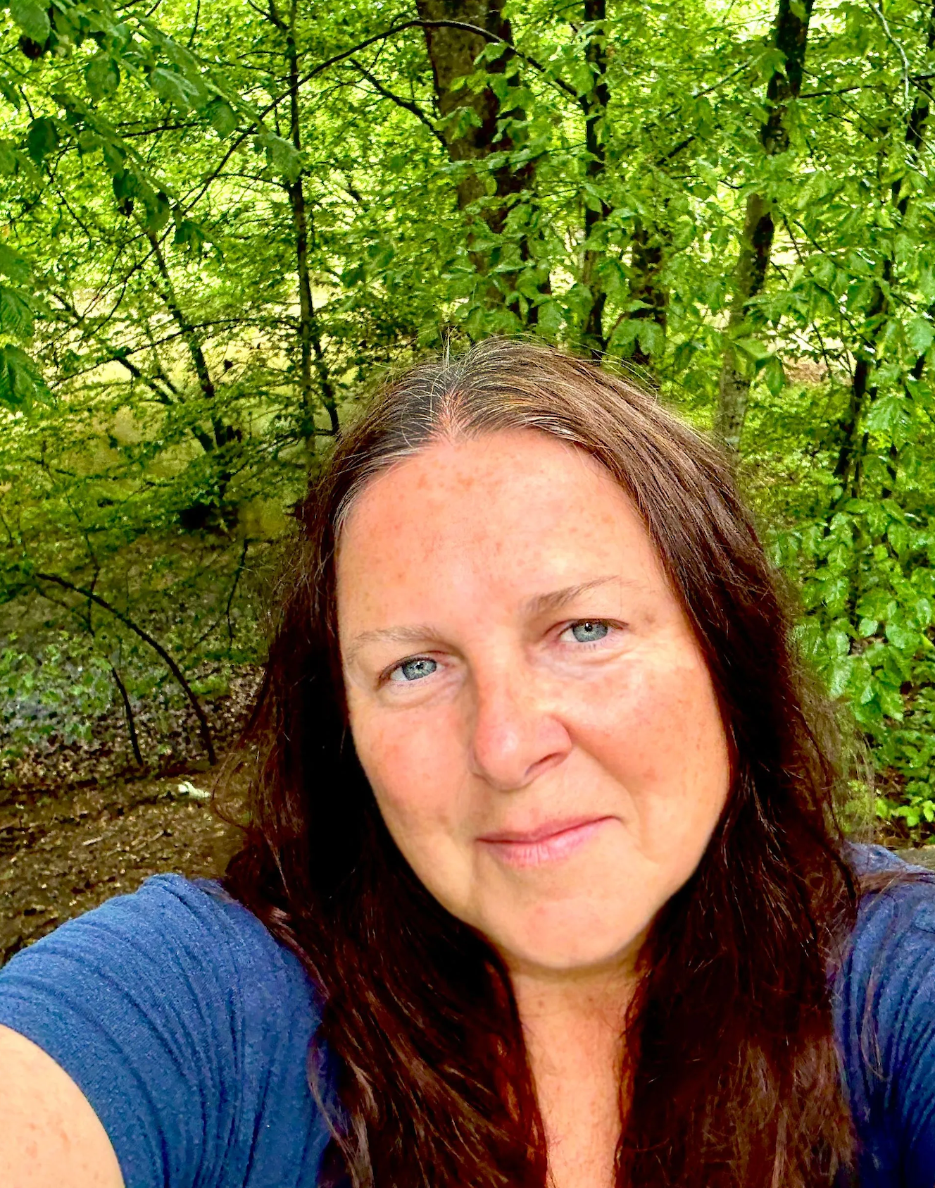 Woman with dark red hair taking a selfie outdoors in a lush green forest setting.