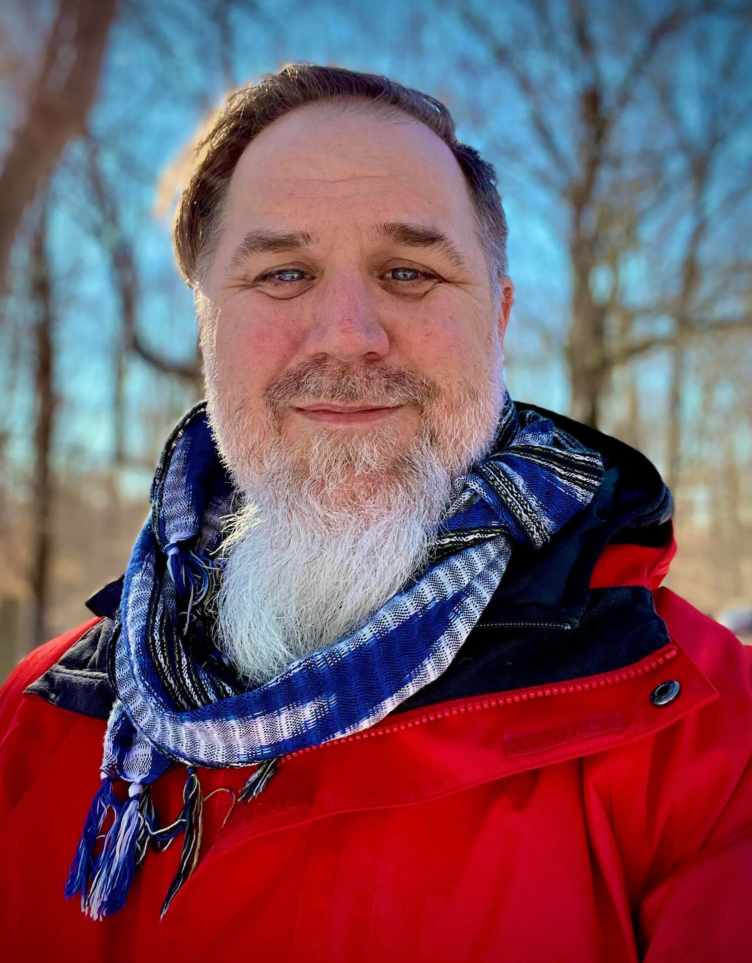 Bearded man wearing a red jacket and blue scarf smiling outdoors in winter with bare trees behind him.