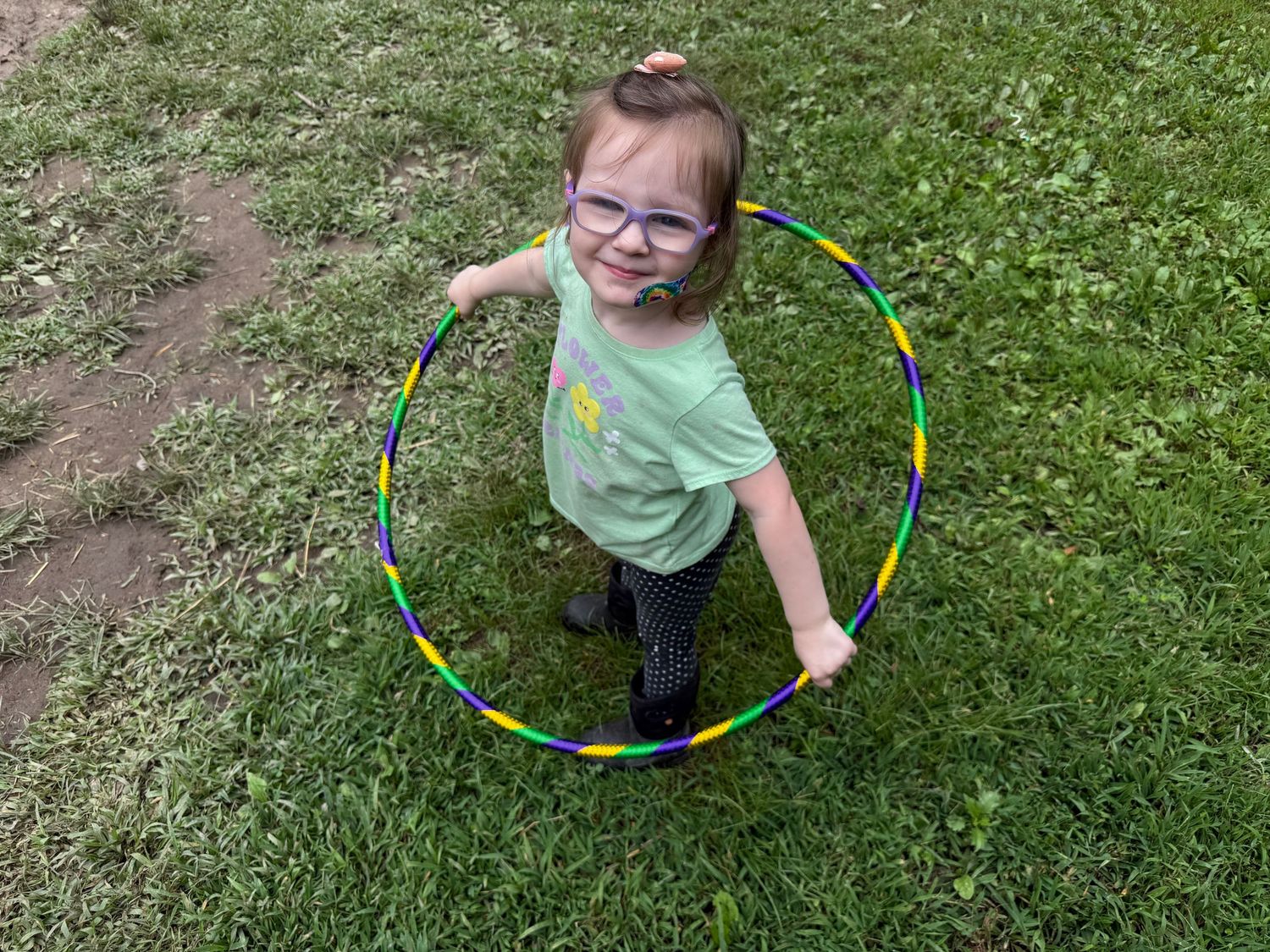 Young girl with glasses holds a colorful rainbow hula hoop while standing on green grass outdoors.