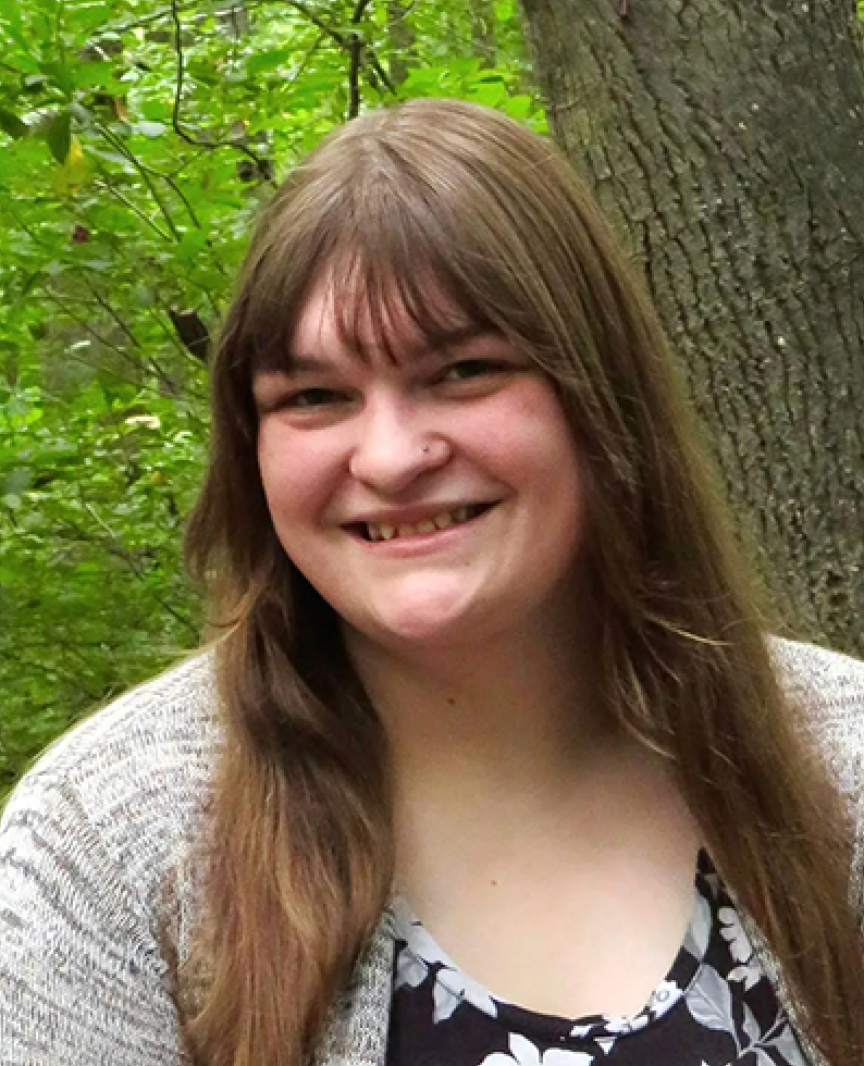 Smiling young woman with long brown hair wearing a floral top and gray cardigan, standing outdoors near trees.