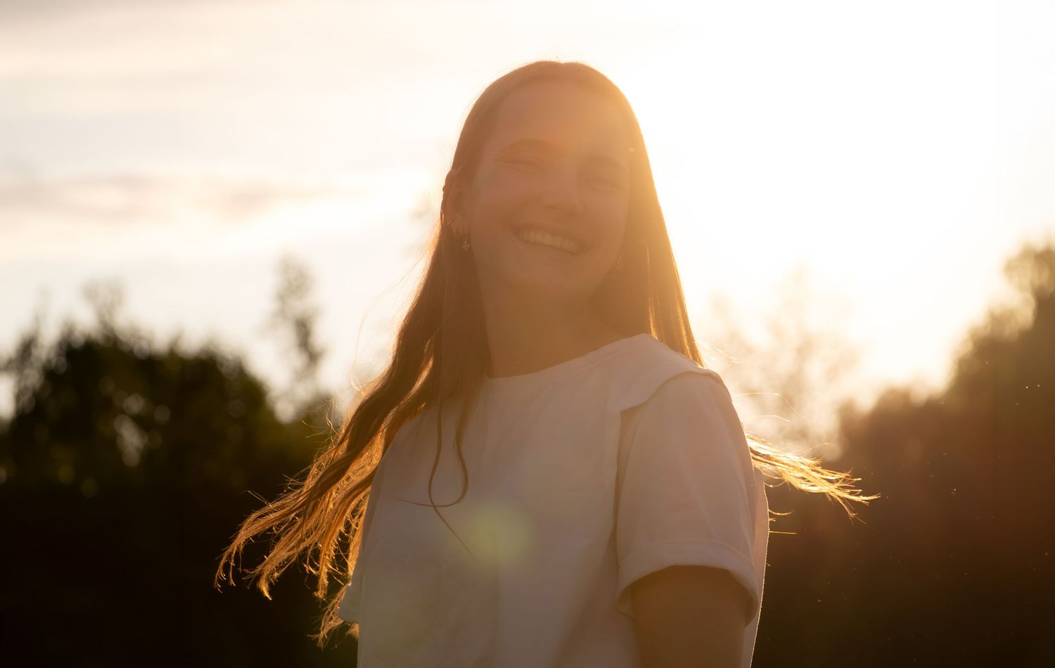 Smiling teenage girl with long hair in white t-shirt, backlit by golden sunset light outdoors.