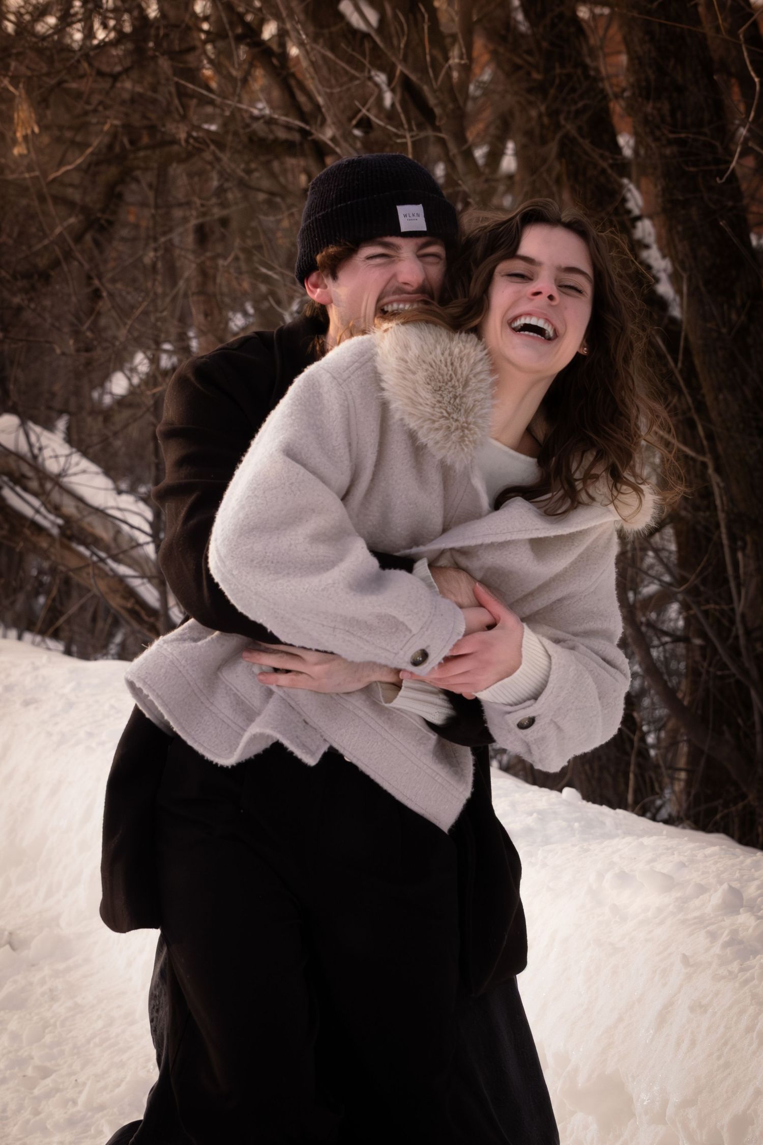 A couple shares a joyful embrace while laughing together in a snowy winter forest setting.