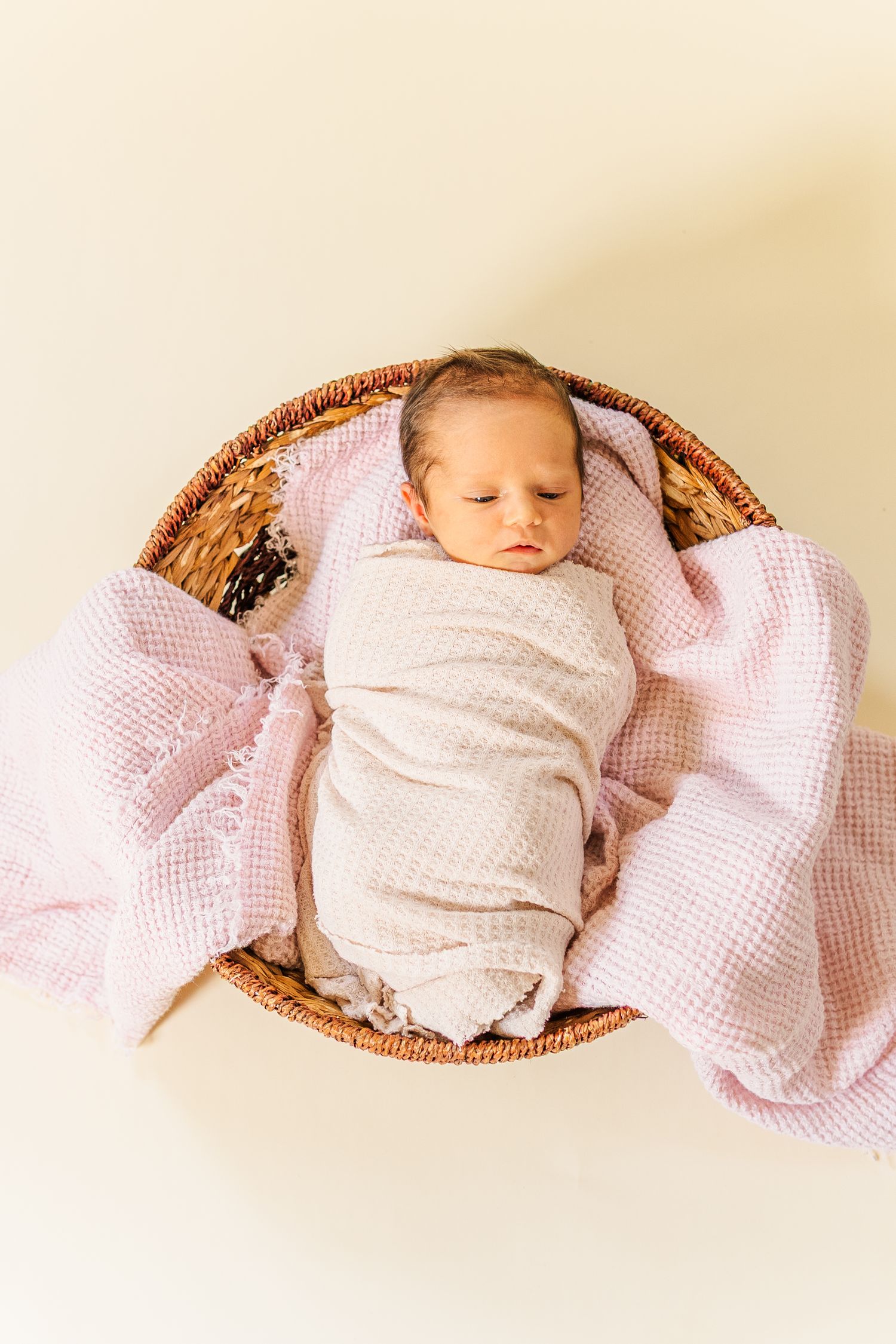 A sleeping newborn baby swaddled in white fabric lies peacefully in a woven basket against a cream background.