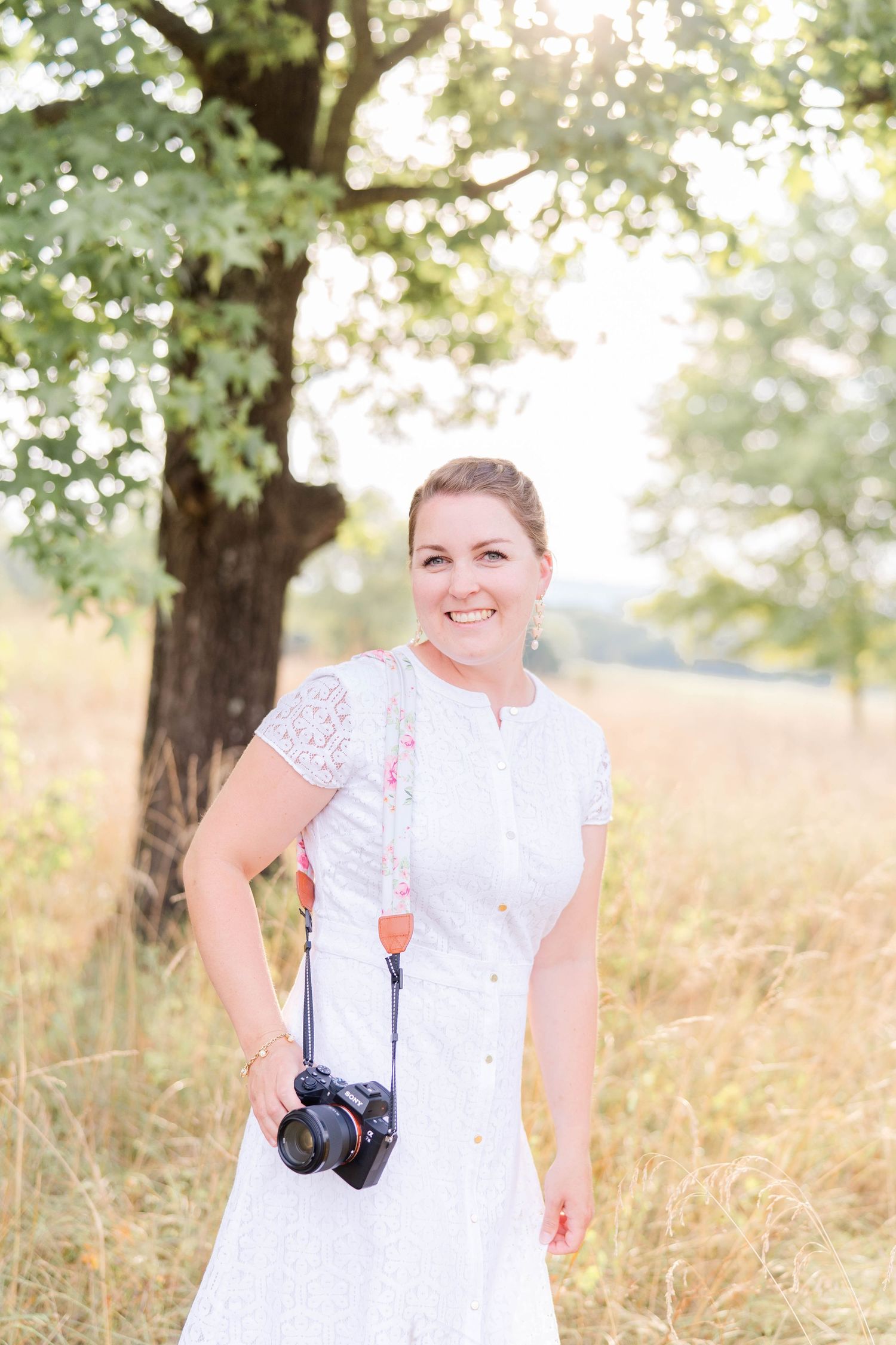 A photographer in a white lace dress stands in a sunlit field with a camera hanging from their shoulder.