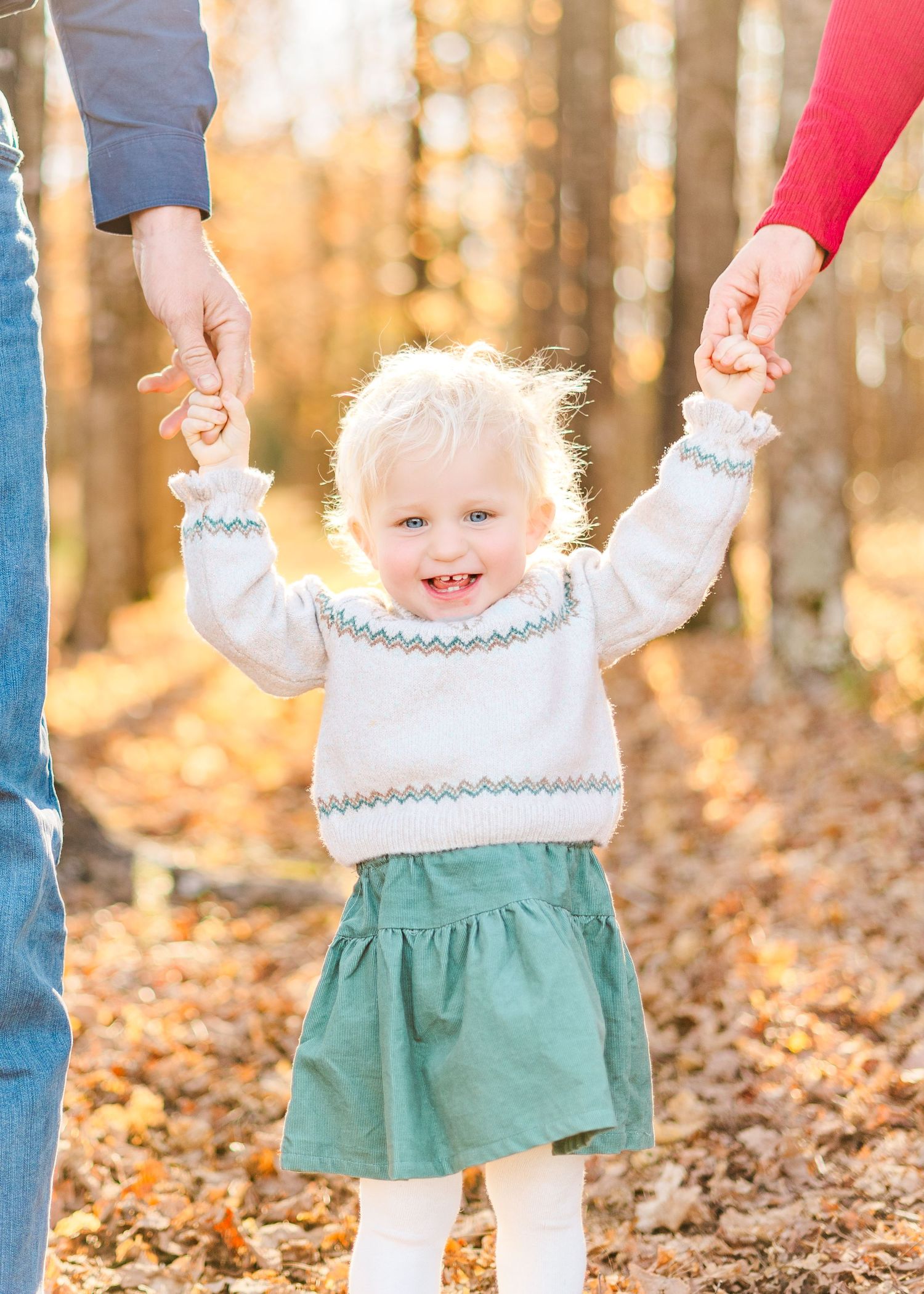 Children in matching white and green dresses walk hand in hand with adults through autumn leaves in a forest.