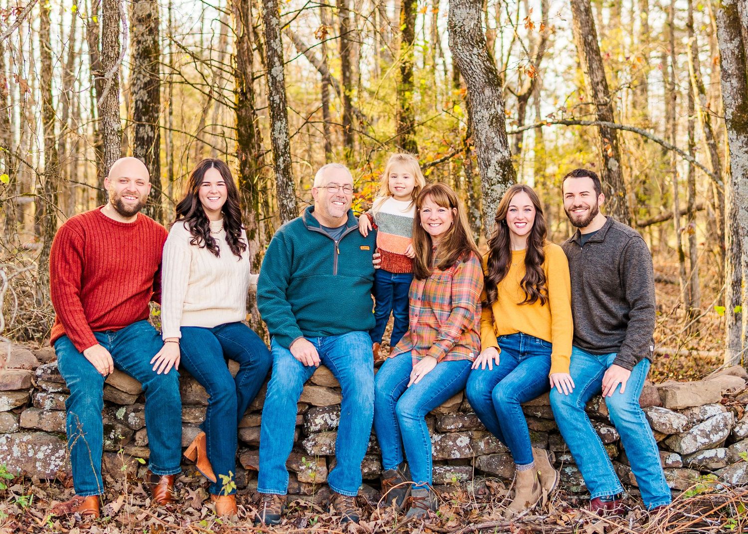 Group of people wearing coordinated denim and colorful tops sit together on a log in an autumn forest setting.