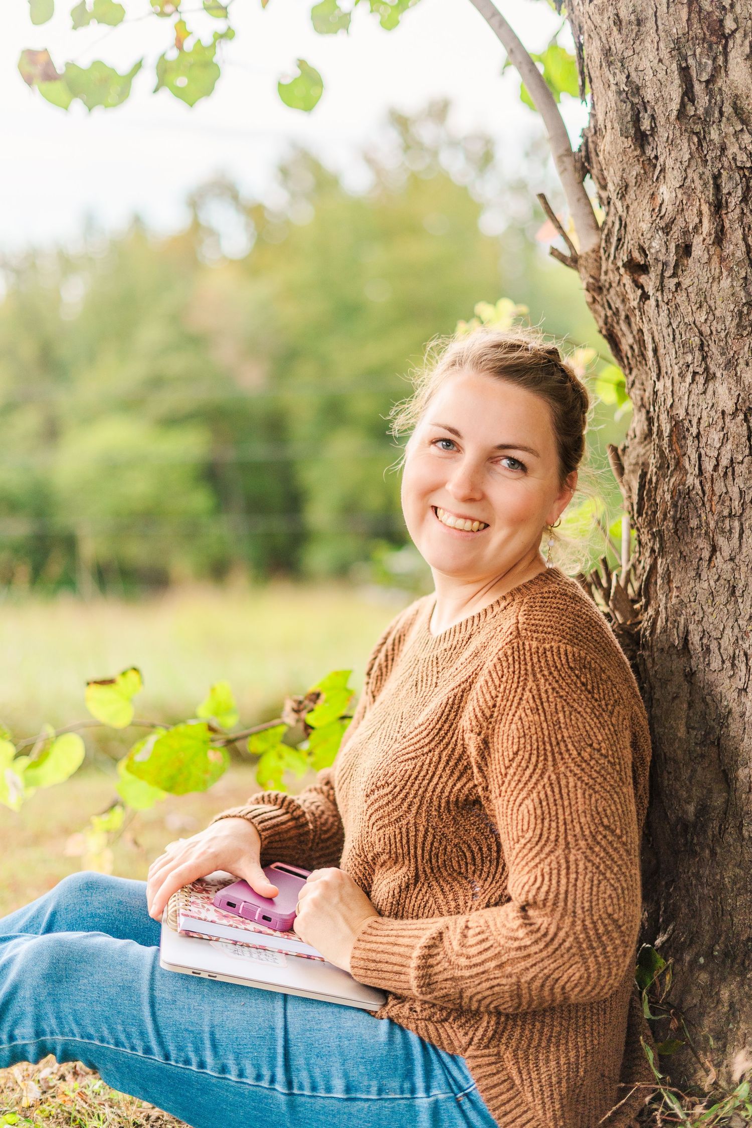 Person in brown knit sweater relaxing against tree trunk while reading book in autumn outdoor setting.