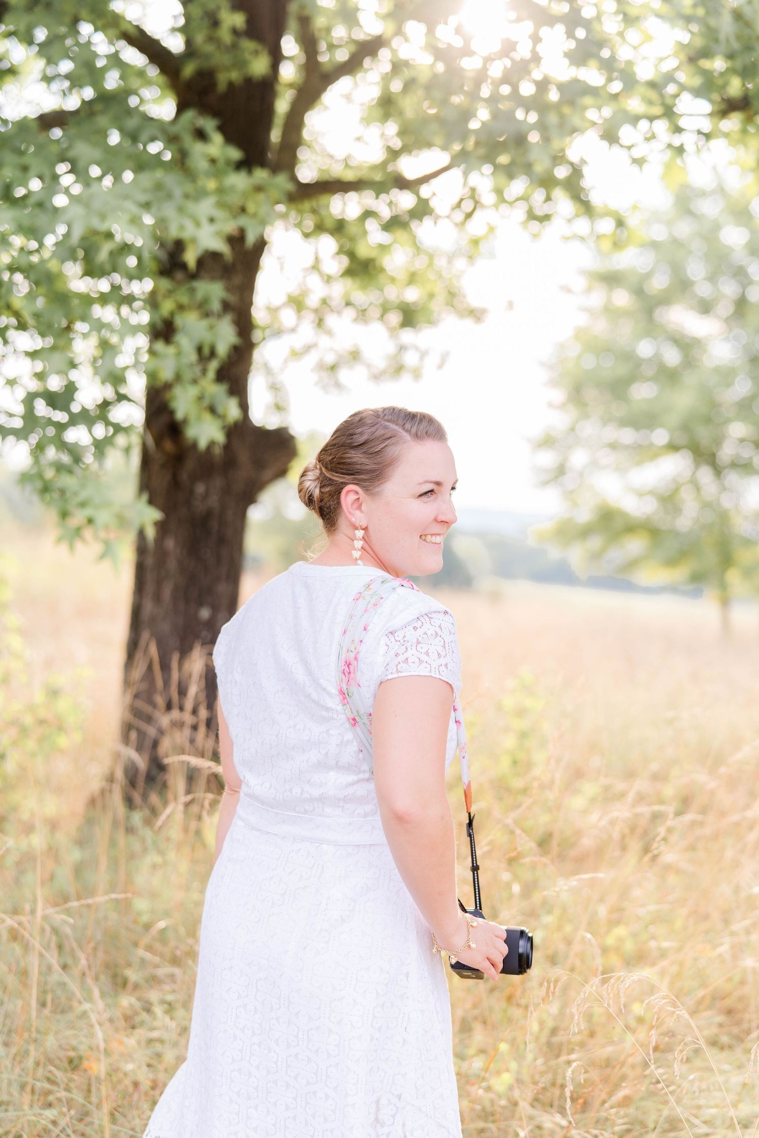 A person in a white lace wedding dress stands in a sunlit meadow near a large tree on a summer day.
