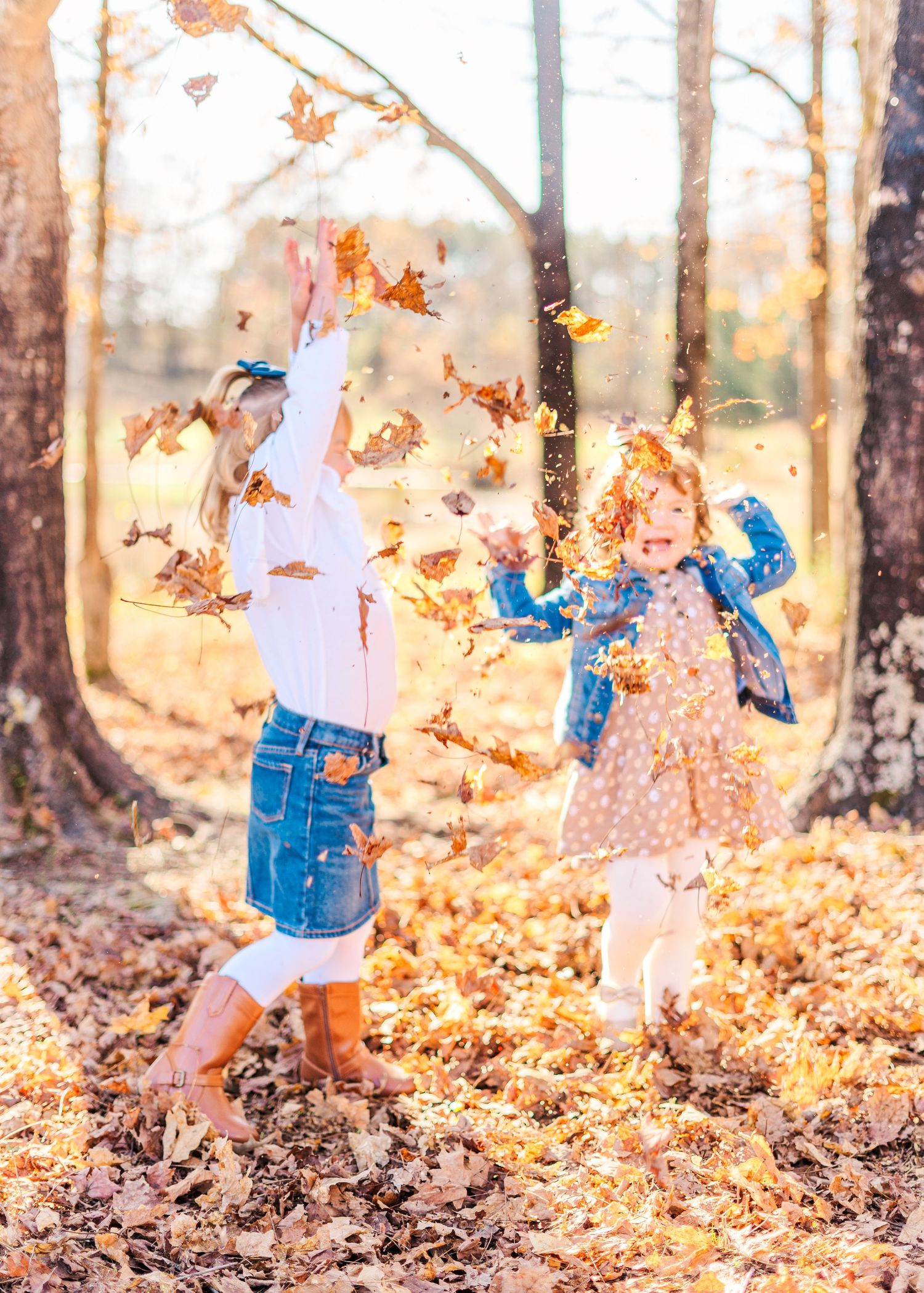 Child in denim and white plays joyfully in a pile of autumn leaves at sunset in the woods.