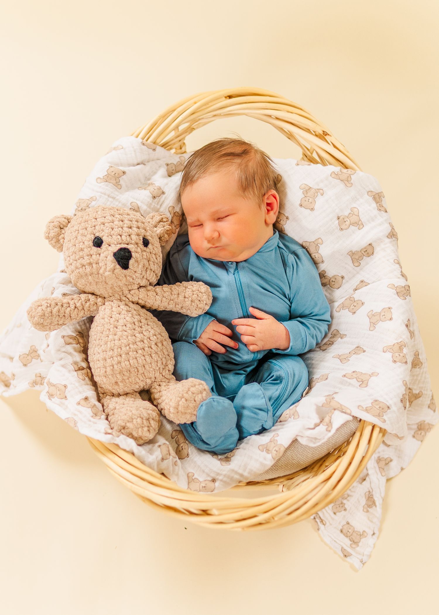 Sleeping baby in blue outfit cuddled with plush teddy bear in woven basket on patterned blanket.