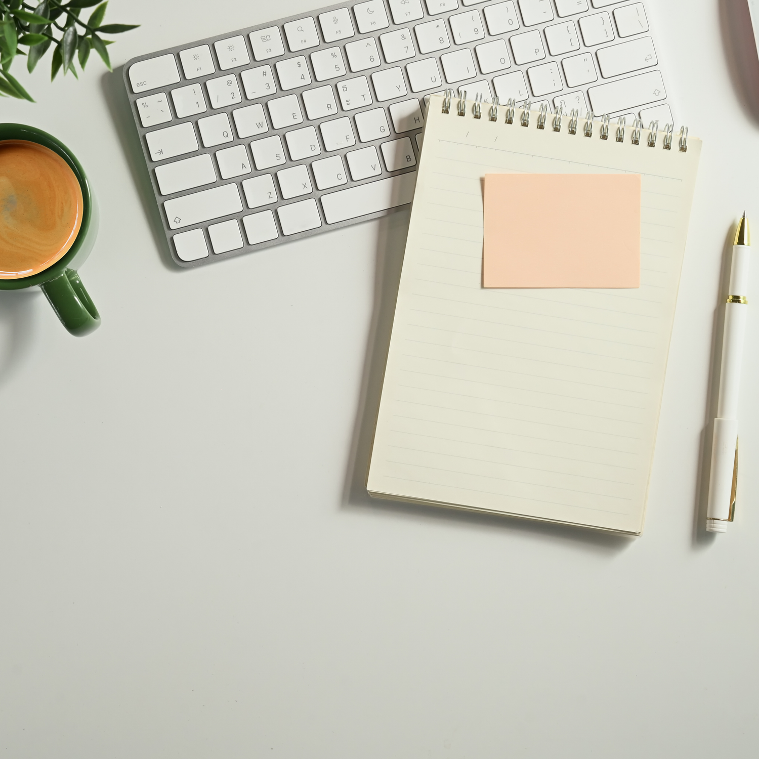 Minimalist workspace with white keyboard, spiral notebook, sticky note, green coffee mug and plant on gray desk.