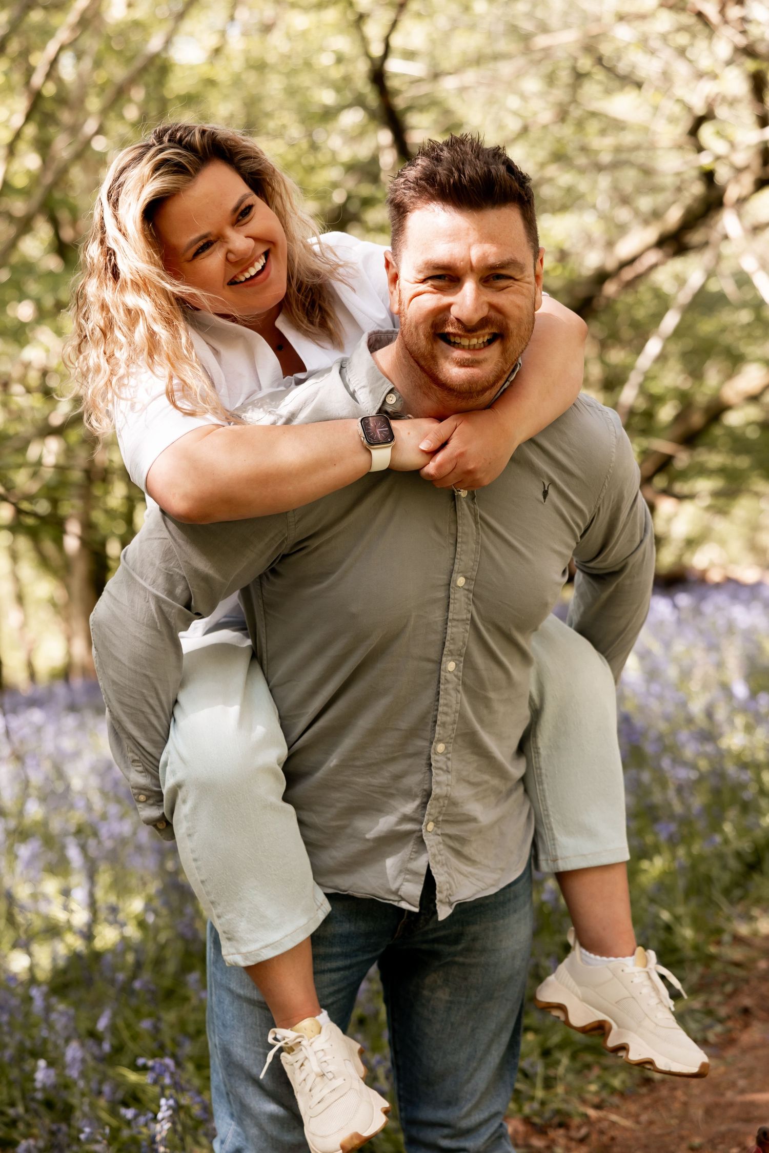 Happy couple laughing in bluebell woods, woman piggyback riding on mans back among spring forest trees.
