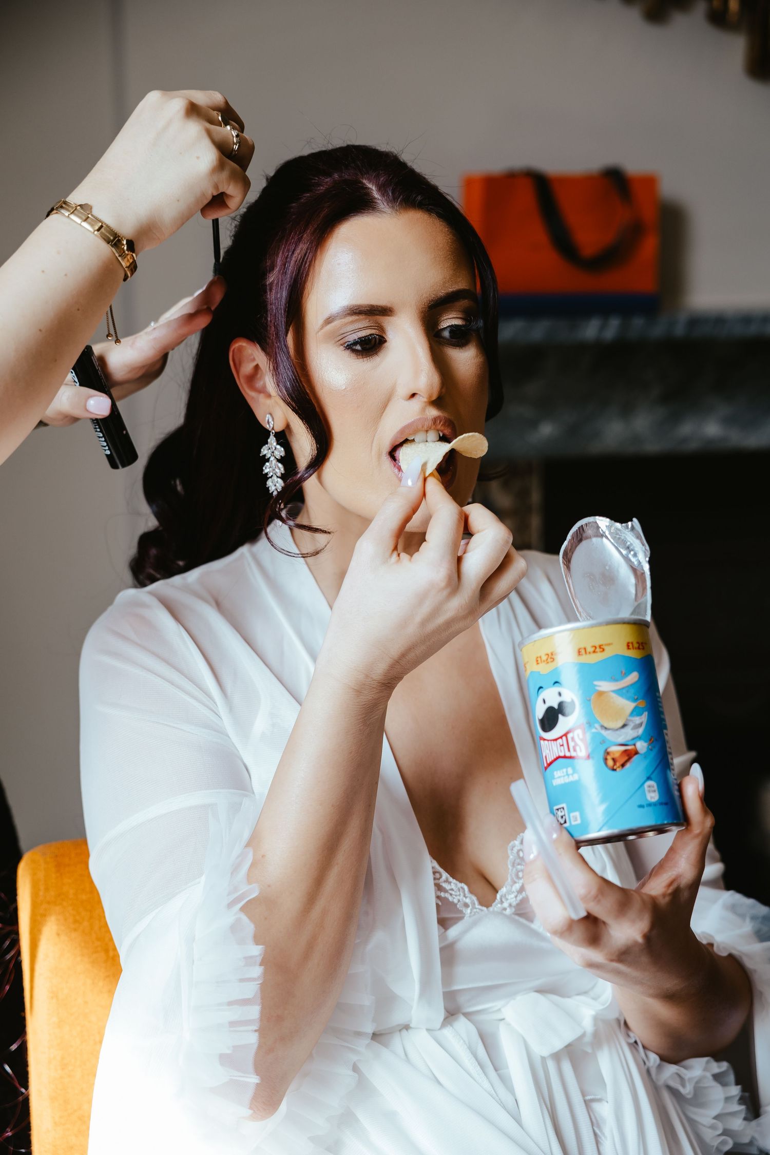 Bride in white robe eating Pringles chips while getting hair done before wedding ceremony.