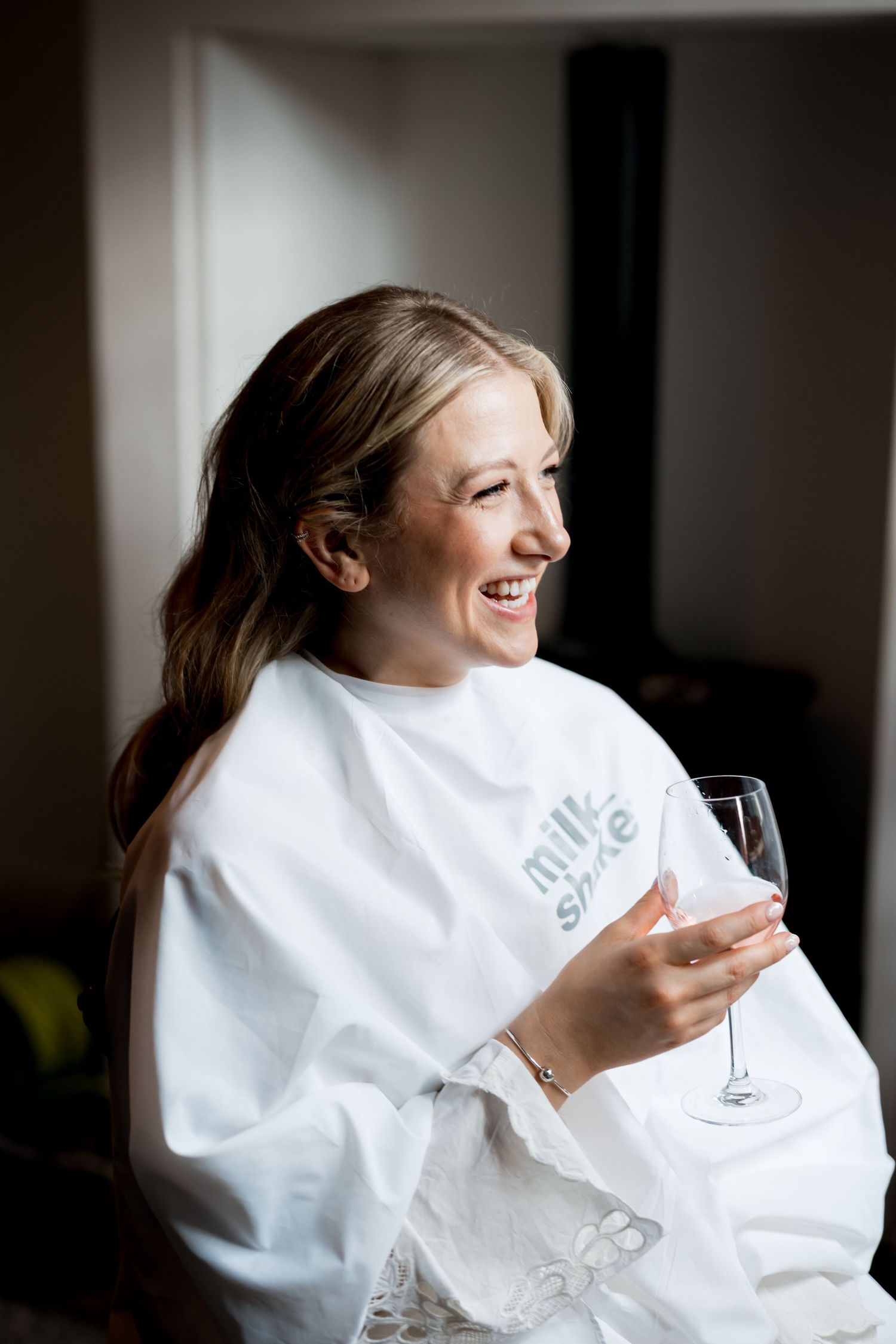 Smiling blonde woman in a white milkshake salon cape holding a wine glass during bridal hair preparation.