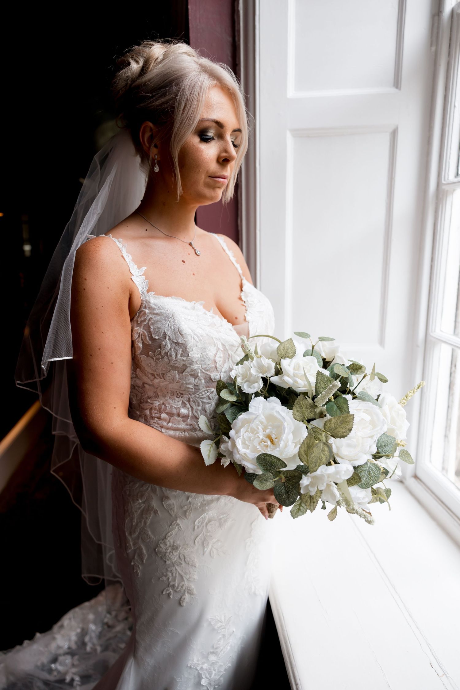 Bridal portrait by window showing detailed white lace dress and white floral bouquet.